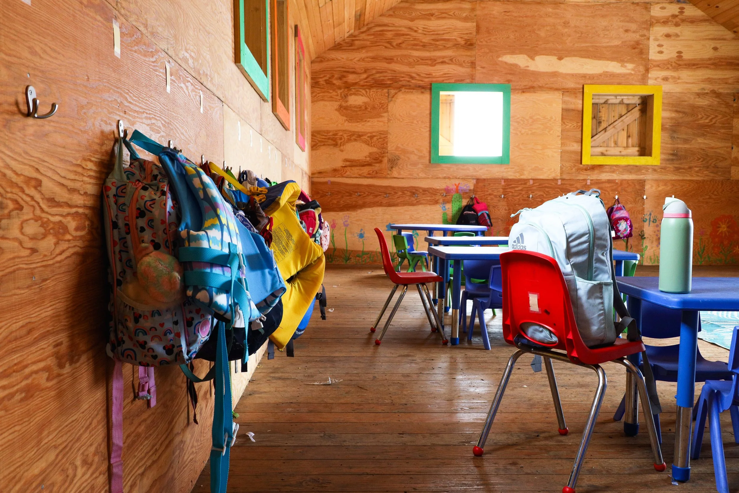 A colorful classroom with backpacks hanging on hooks, tables with plastic chairs, and bright windows on wooden walls decorated with children's artwork.