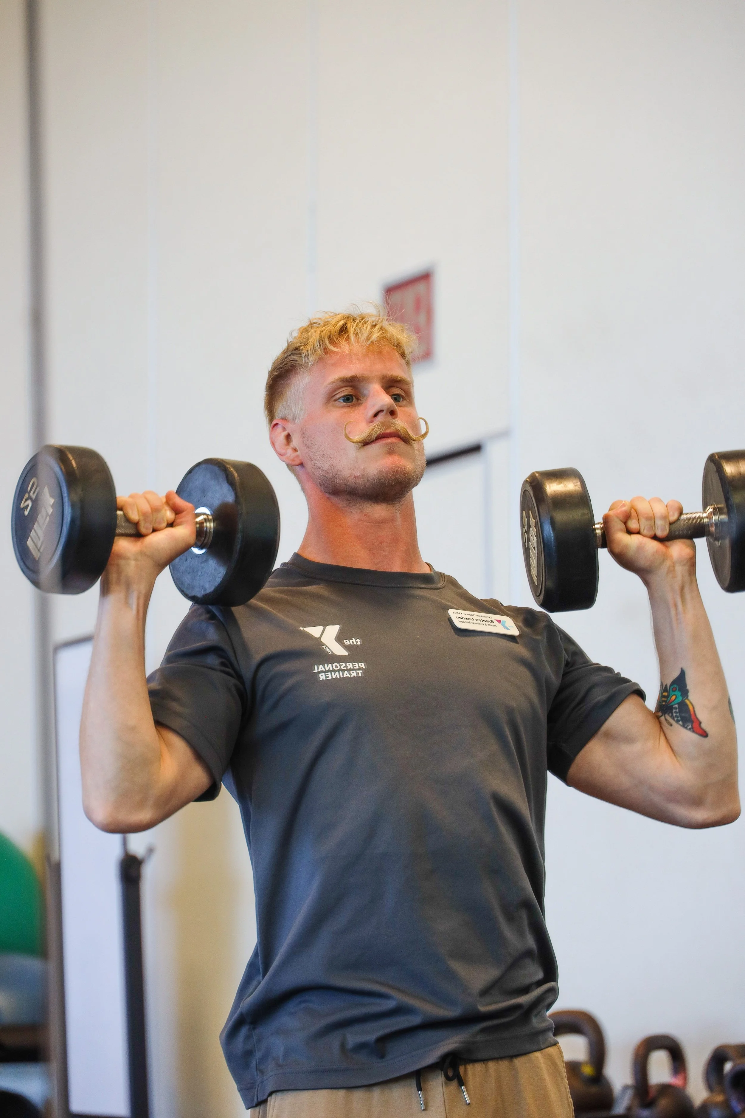A man with blonde hair, a mustache, and wearing a dark gray t-shirt is lifting dumbbells at a gym.