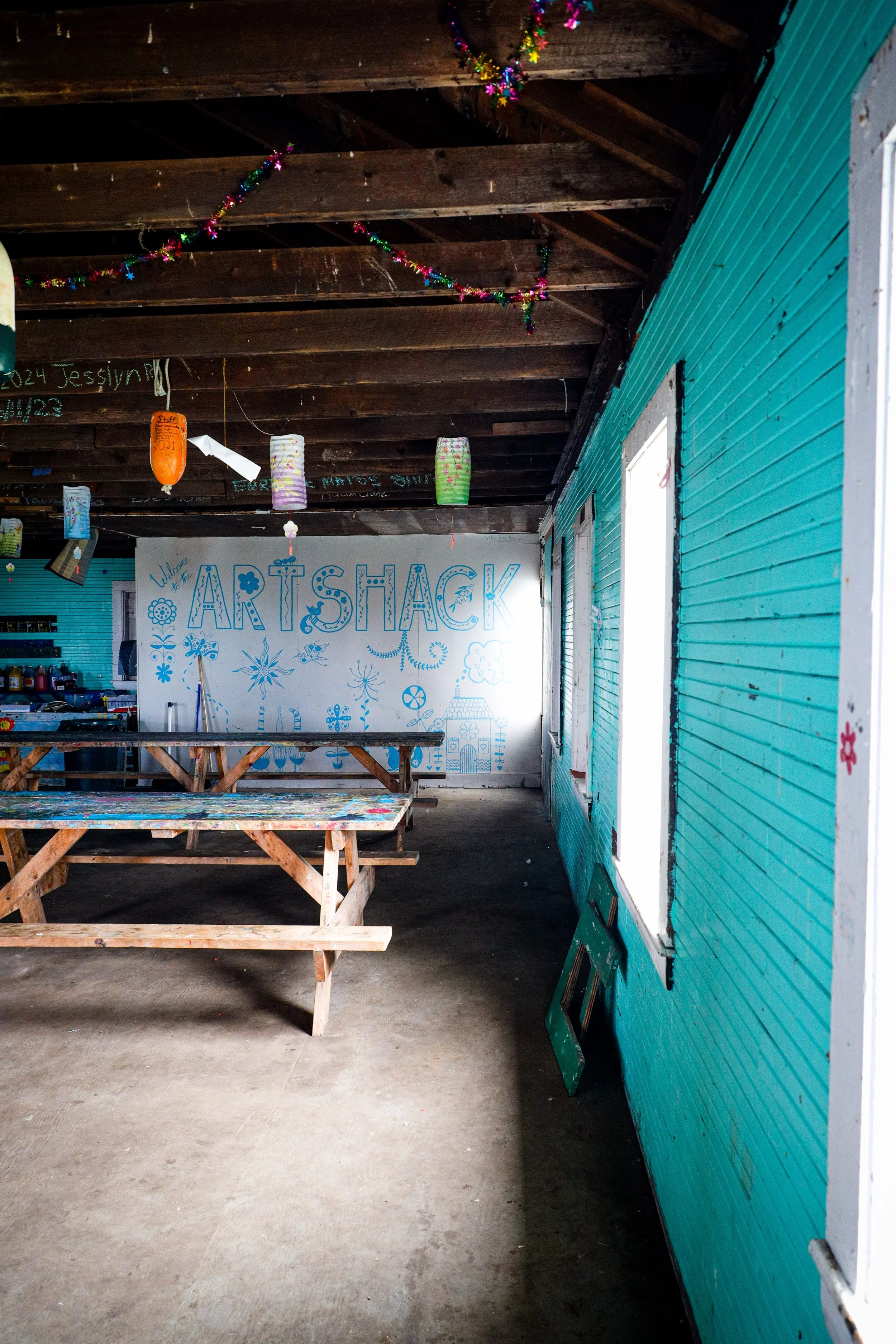 Interior of an art space called Art Shack with decorate tables, turquoise siding, and a white wall with blue artwork and the words "Welcome ART SHACK."