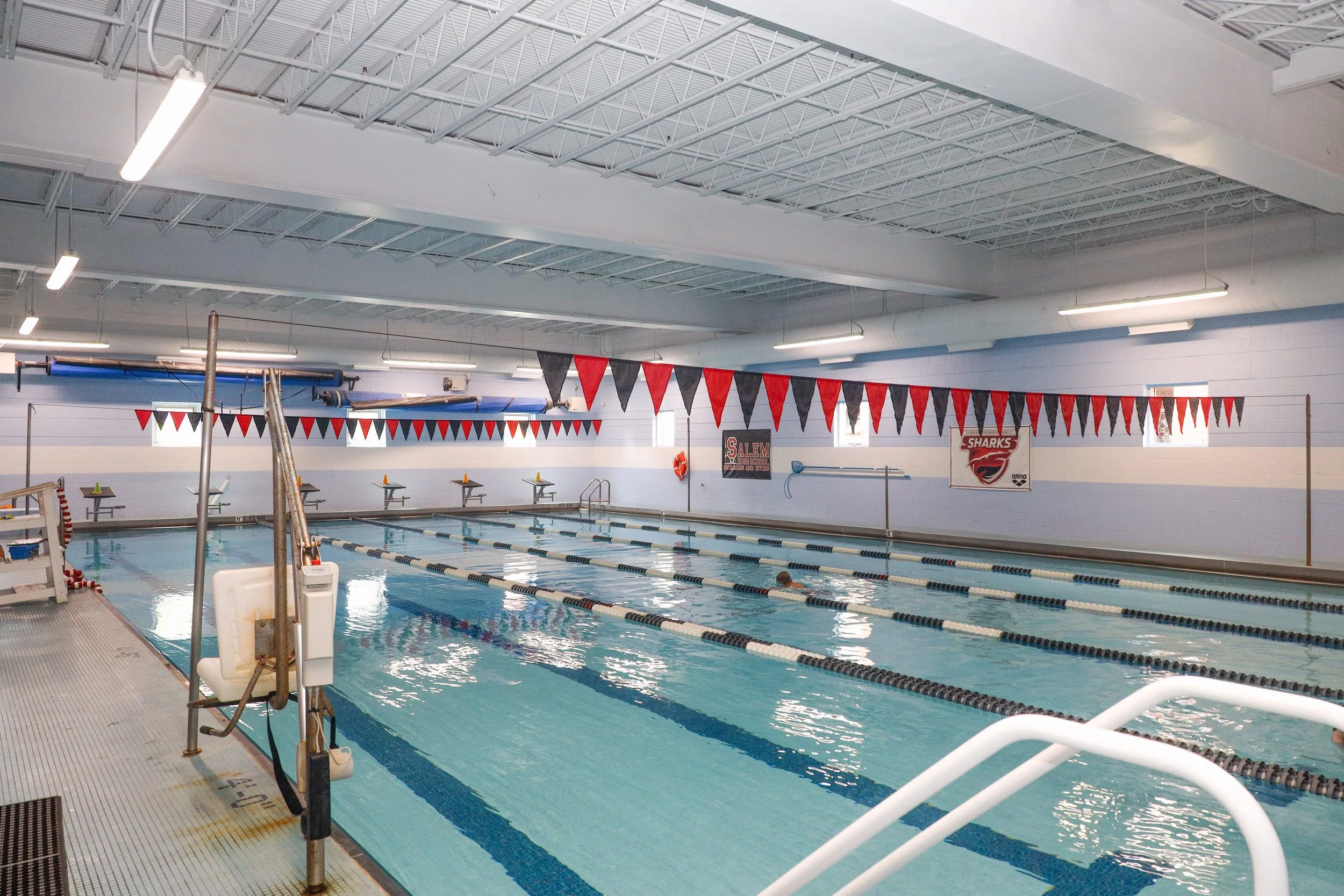 Indoor swimming pool with red, black, and white triangular flags hanging from the ceiling. Pool lanes separated by lane lines, some swimmers in the water. Starting blocks at the far end. Blue and white walls with swim team banners.