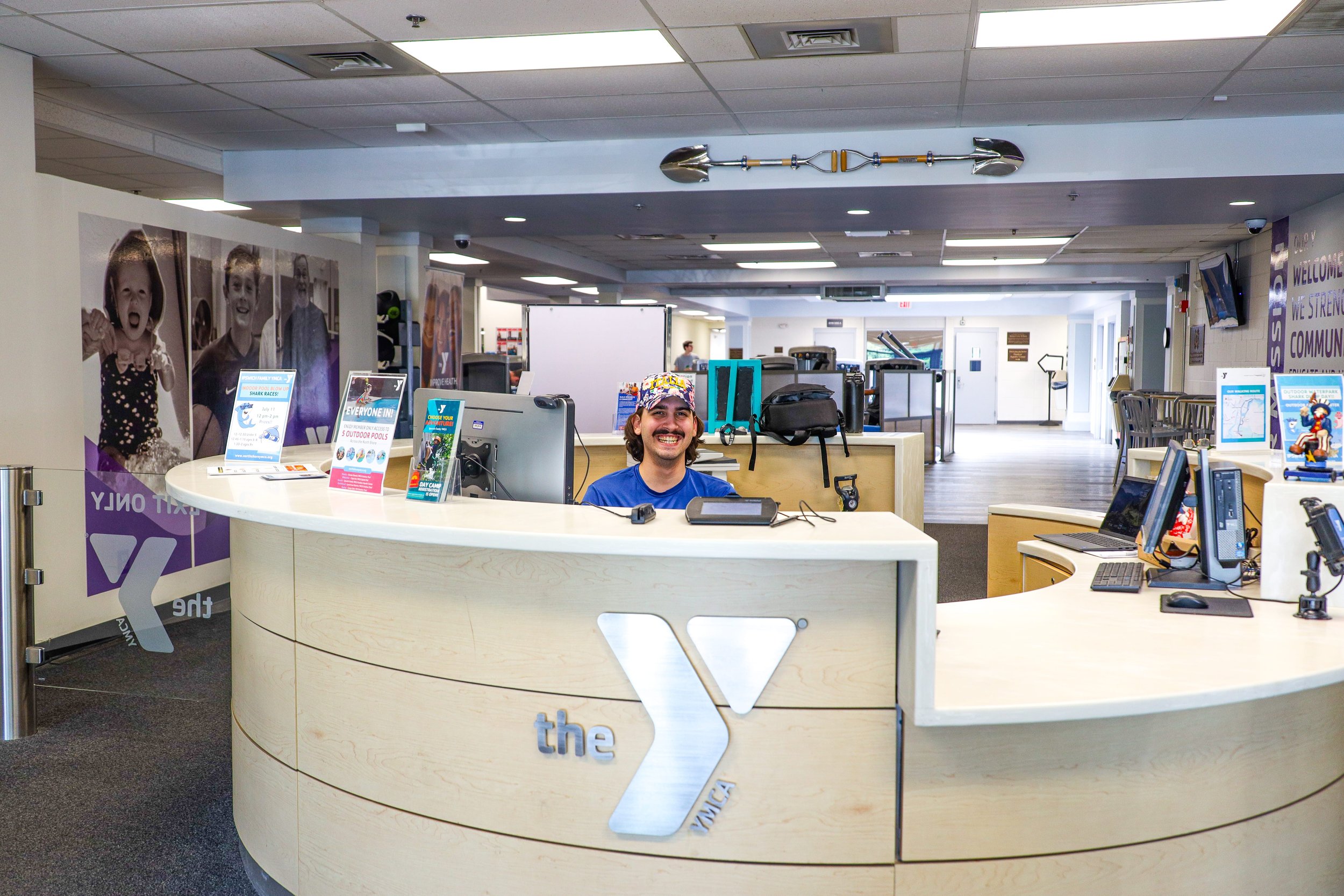 Front desk receptionist smiling at YMCA front desk with computer monitors and brochures, in a large well-lit community center or gymnasium.