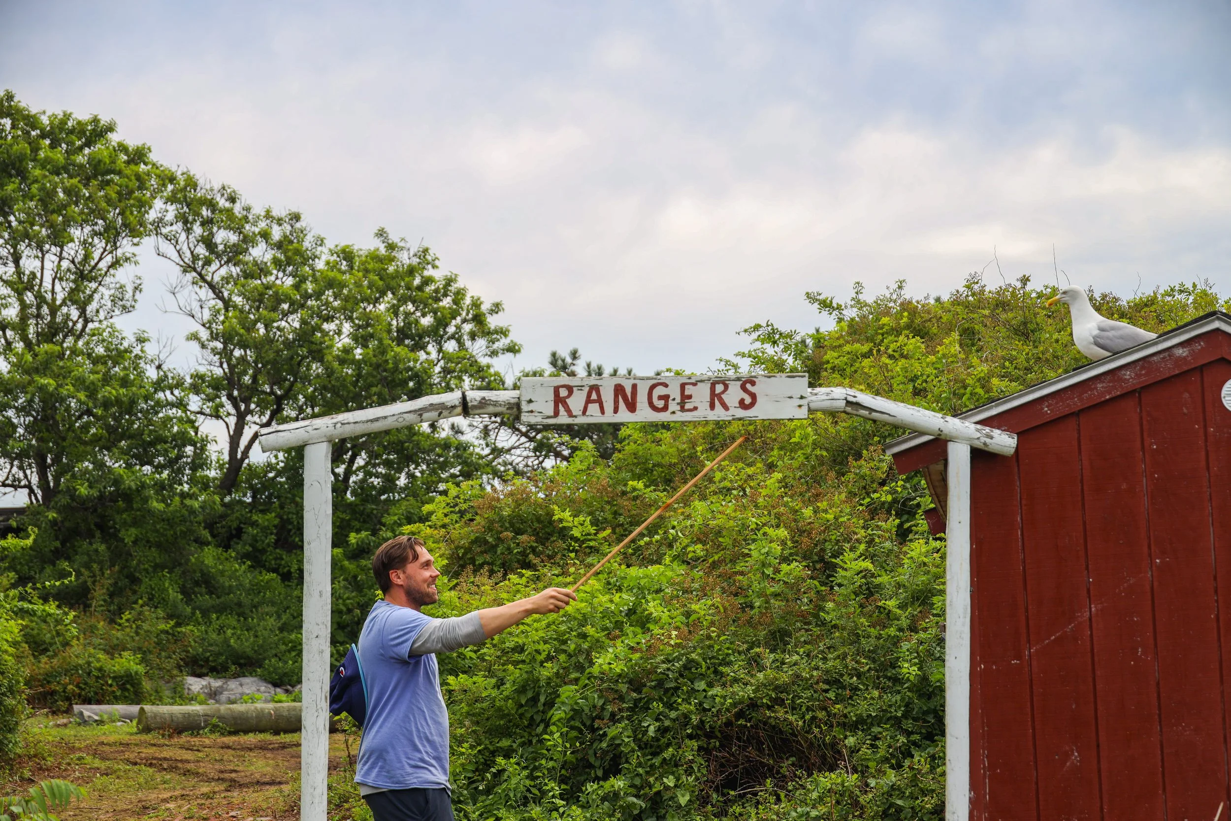 A man standing outdoors, smiling and pointing at a sign that says 'RANGERS' with a stick. There are green trees in the background, a small red wooden structure on the right, and a seagull perched on its roof.