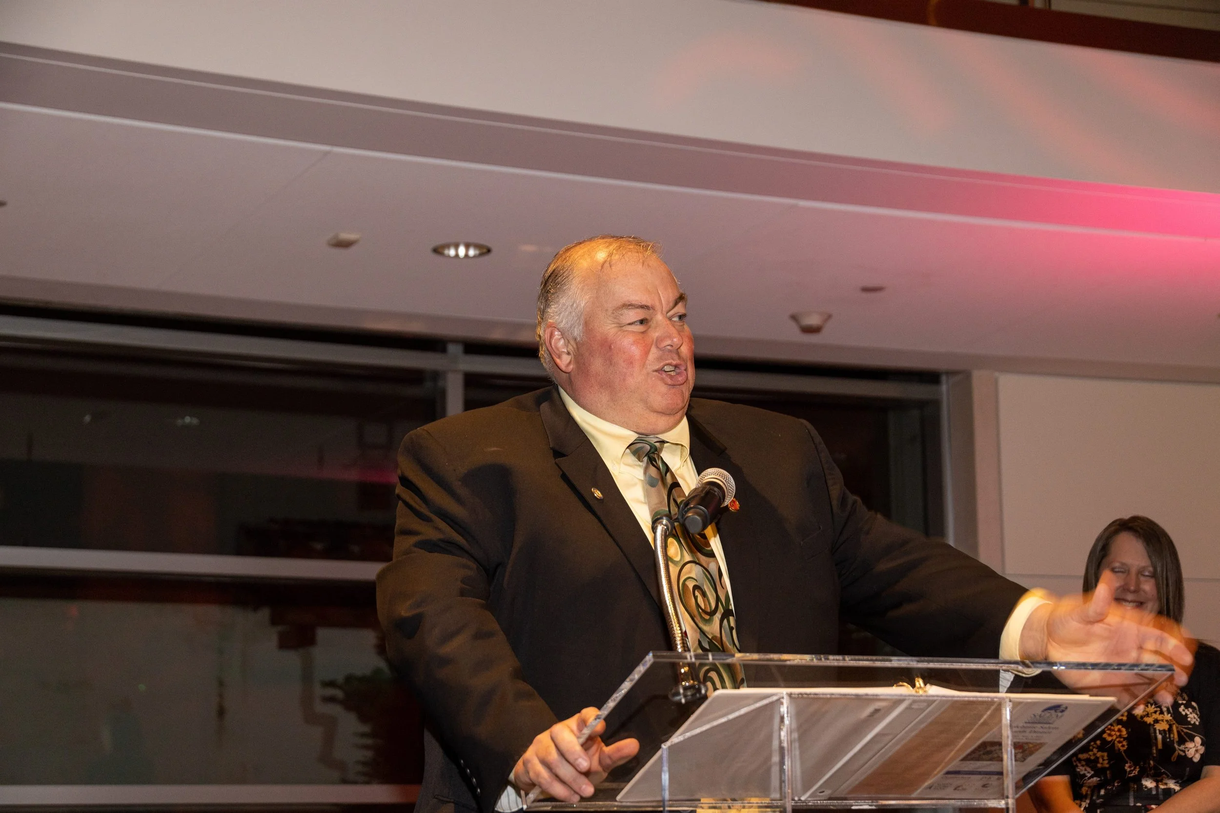 A man in a black suit and patterned tie speaking at a clear podium with a microphone, with a woman smiling in the background.