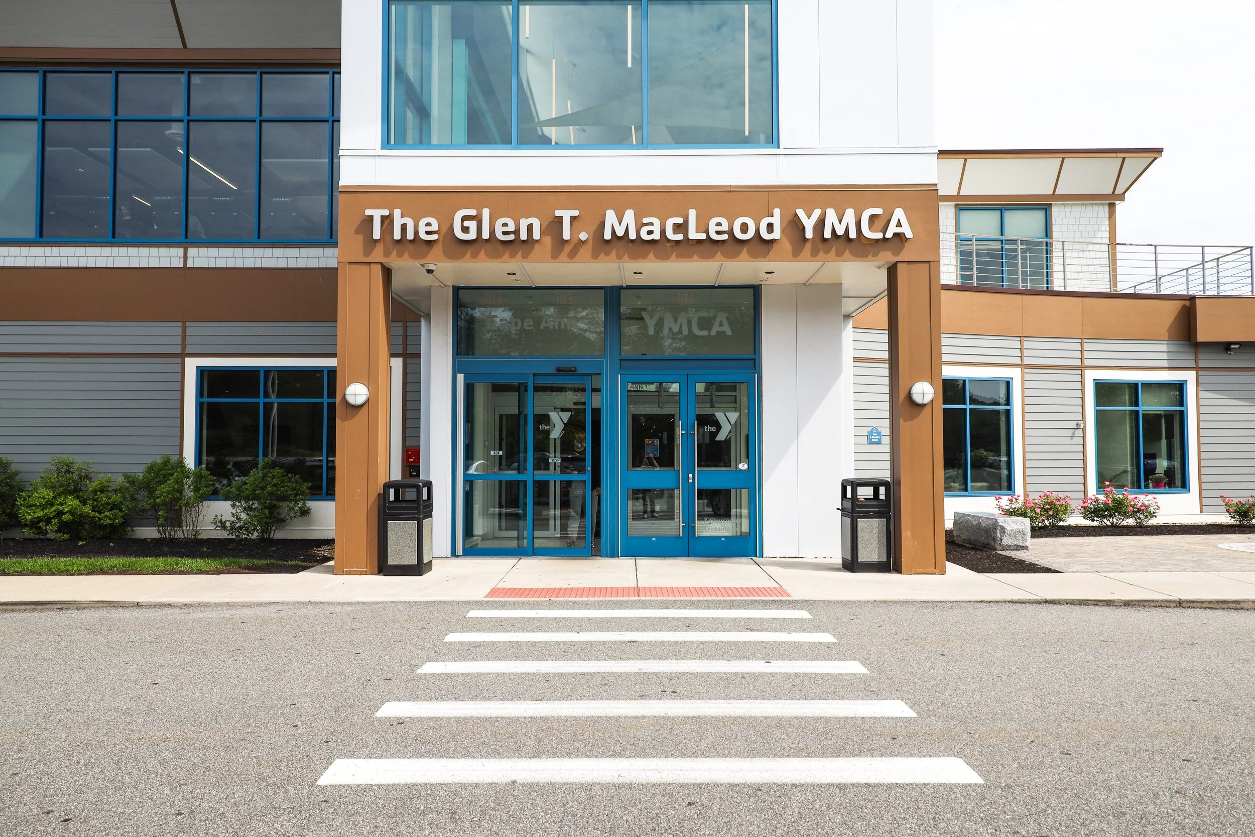 Front entrance of The Glen T. MacLeod YMCA with blue double glass doors, white sign with brown border reading the YMCA, and pedestrian crosswalk in the foreground.