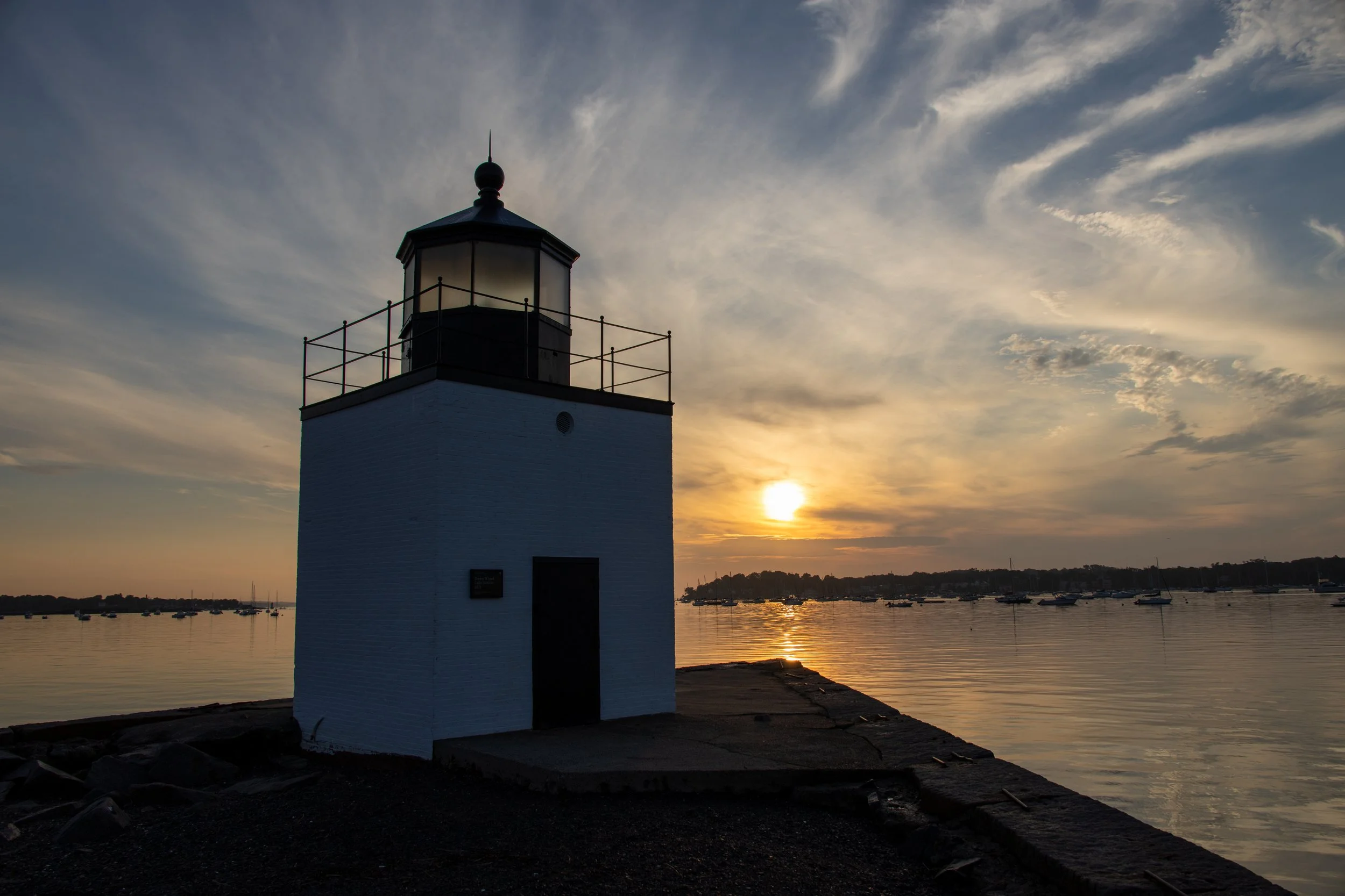 A lighthouse on a pier at sunset, with a cloudy sky and boats in the water in the background.