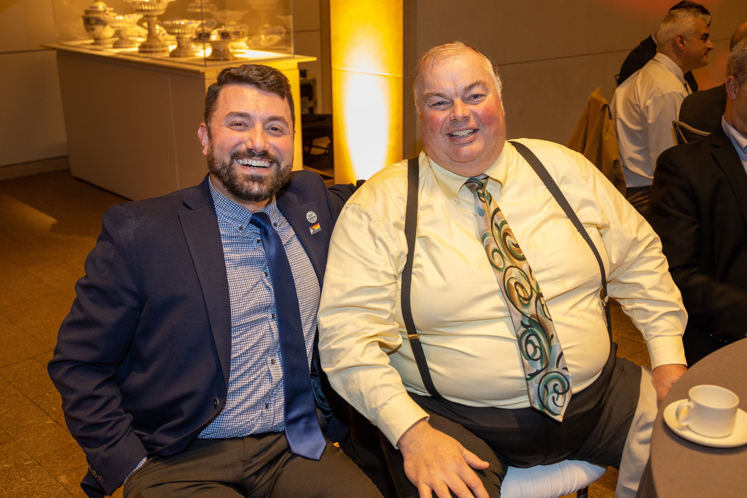 Two men sitting at a table, smiling, dressed in business attire, with one wearing a rainbow pin and the other with suspenders, in a formal event setting.