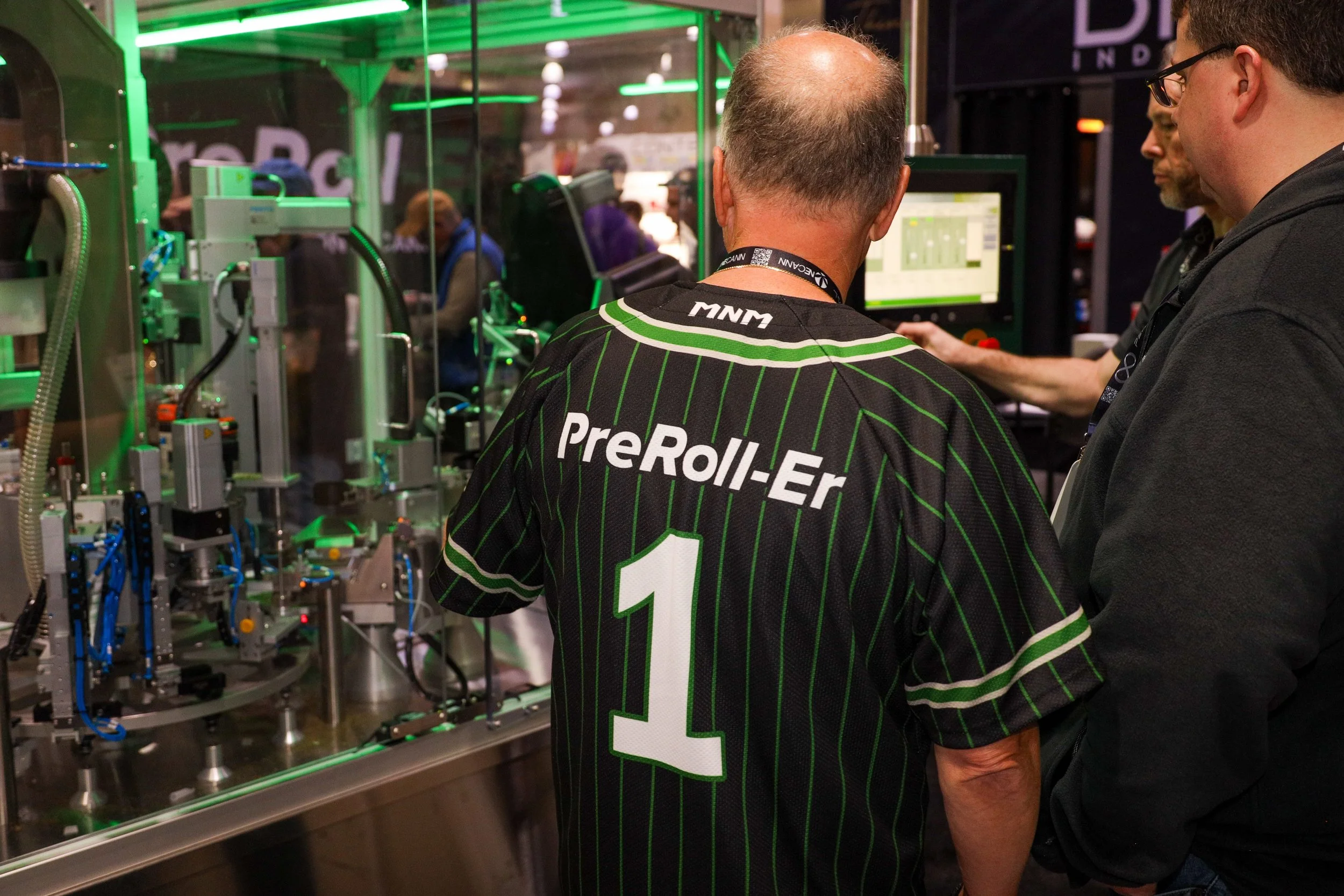 A man wearing a black jersey with green pinstripes and the number one on the back, supporting the brand PreRoll-Er, standing in front of a machinery display at a trade show or exhibition, with two other men observing and discussing the equipment.