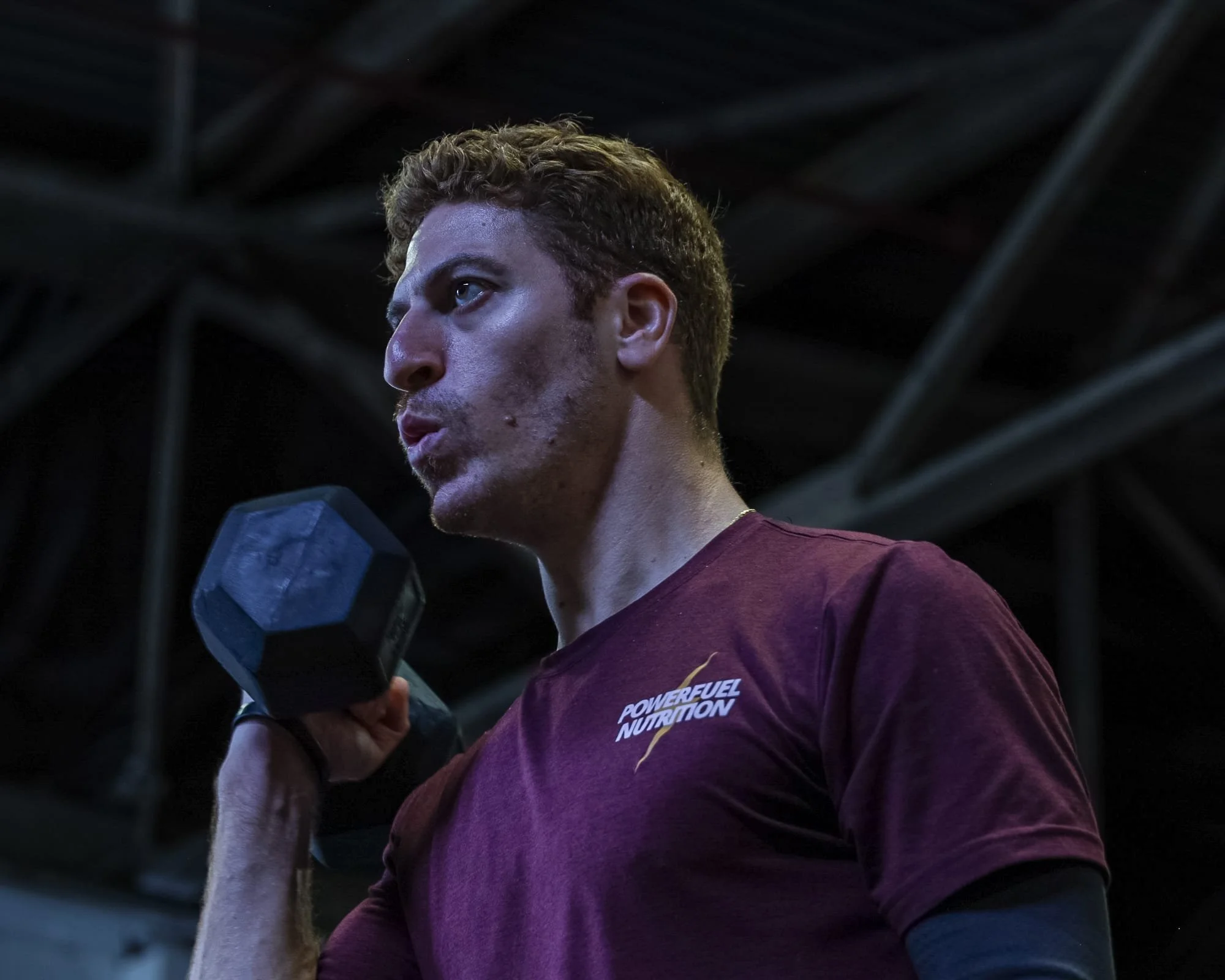 A man lifting a dumbbell in a gym, wearing a maroon shirt with 'Powerful Nutrition' written on it, against a dark industrial background.