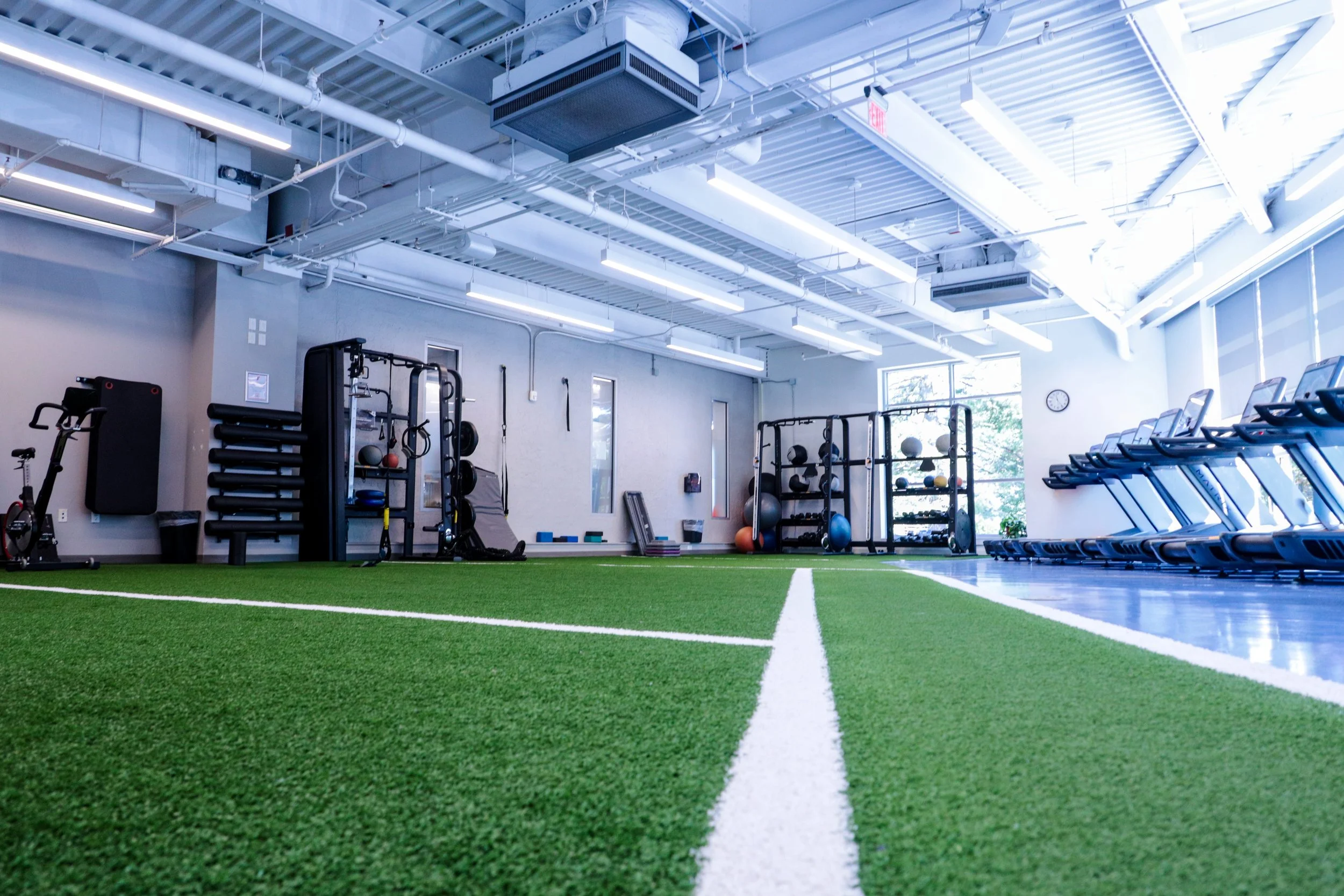 Empty indoor gym with artificial turf, cardio machines on the right, weight racks and fitness equipment along the back wall, large windows letting in natural light, and a clock on the wall.