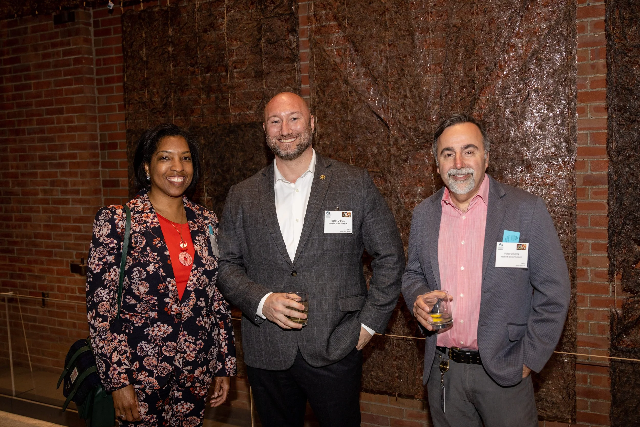 Three people at a social event standing in front of a brick wall with a textured dark brown art piece. The woman on the left is wearing a floral blazer and a red top, the man in the middle is in a gray plaid suit holding a drink, and the man on the right is in a gray blazer with a pink shirt also holding a drink. They are all smiling.