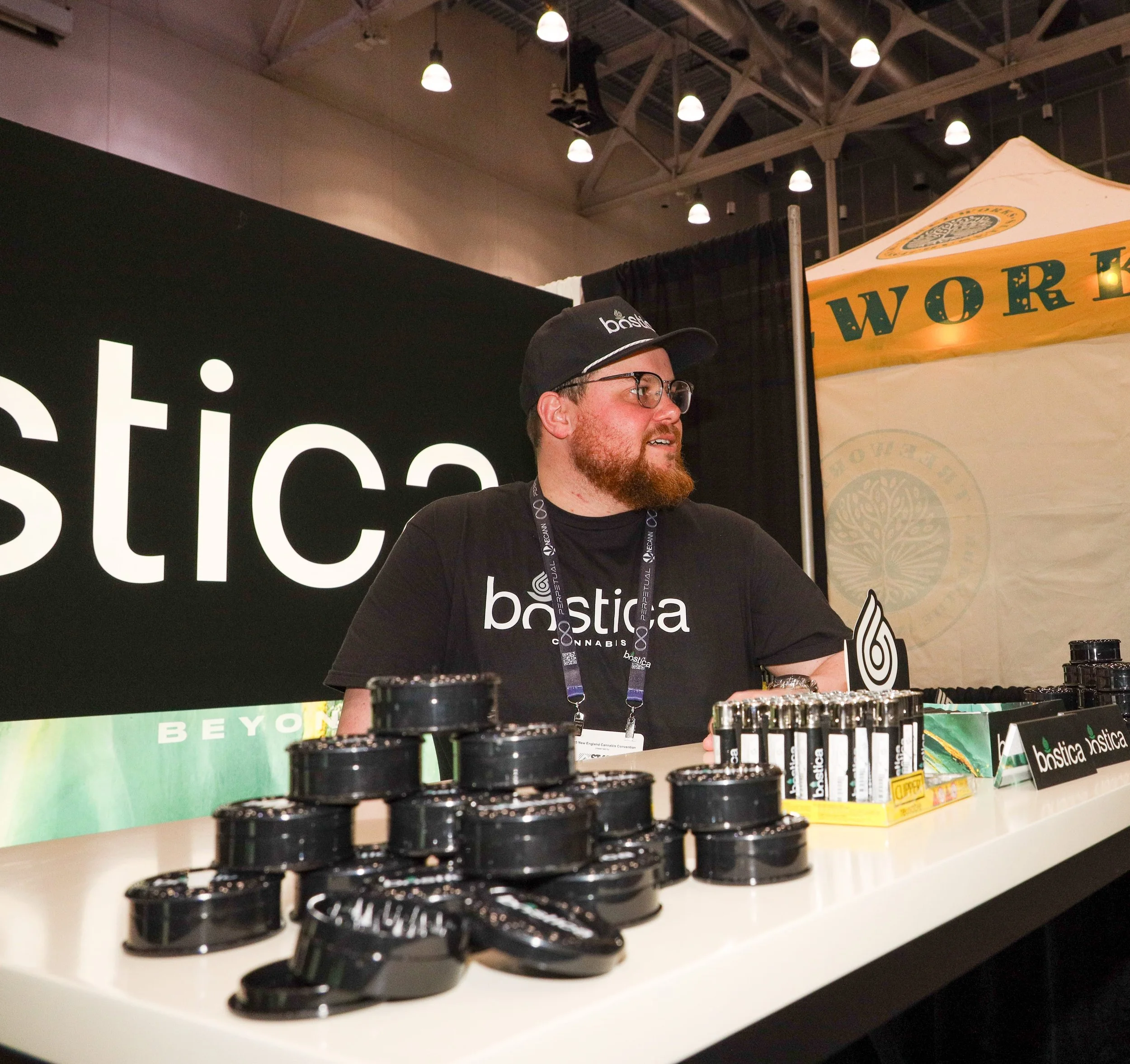 A man with glasses, a beard, and a black cap and t-shirt stands behind a table with stacks of black containers and products, at a booth with black and yellow banners. The booth has the text 'bostica' on the banner and shirt.