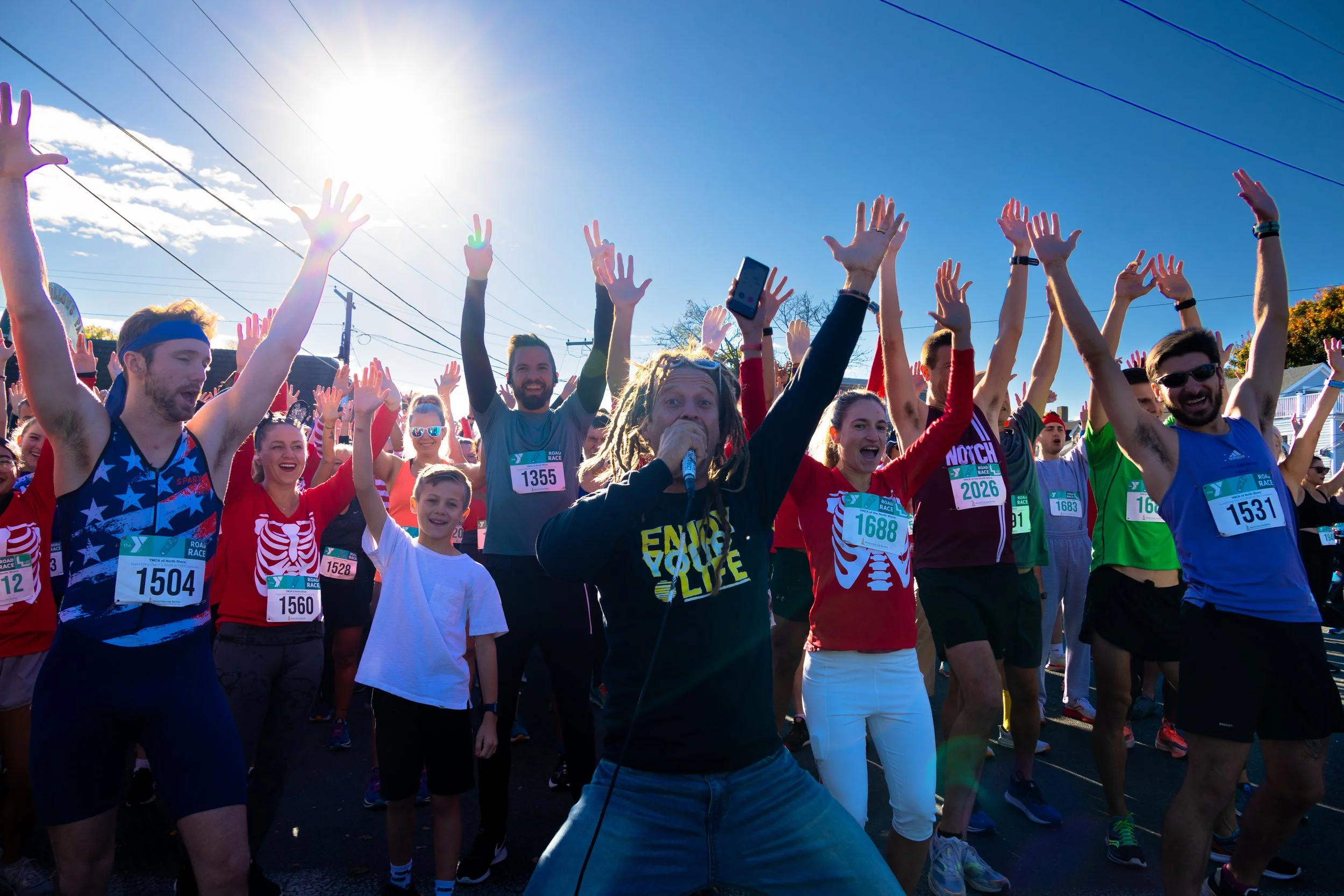 Crowd of runners at the starting line of a race, some with arms raised, with a person with dreadlocks and a microphone leading cheers in the foreground, under a clear blue sky with the sun shining.