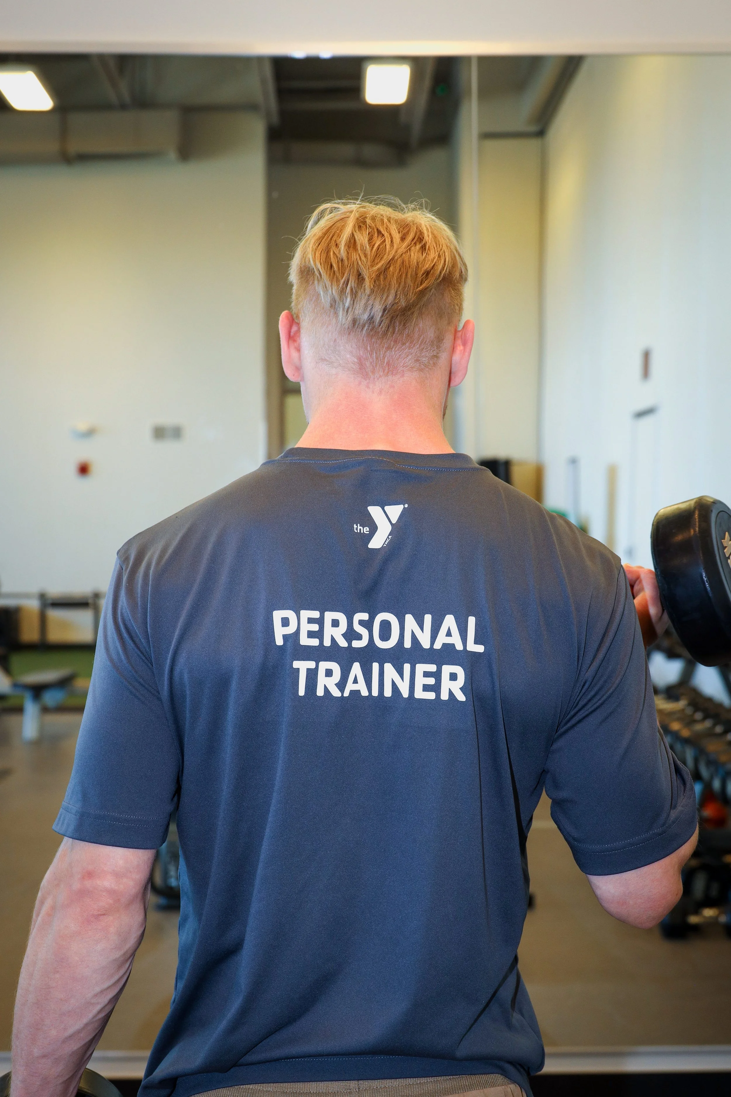 Back view of a man wearing a dark gray shirt with "Personal Trainer" written on it, holding a dumbbell in a gym.