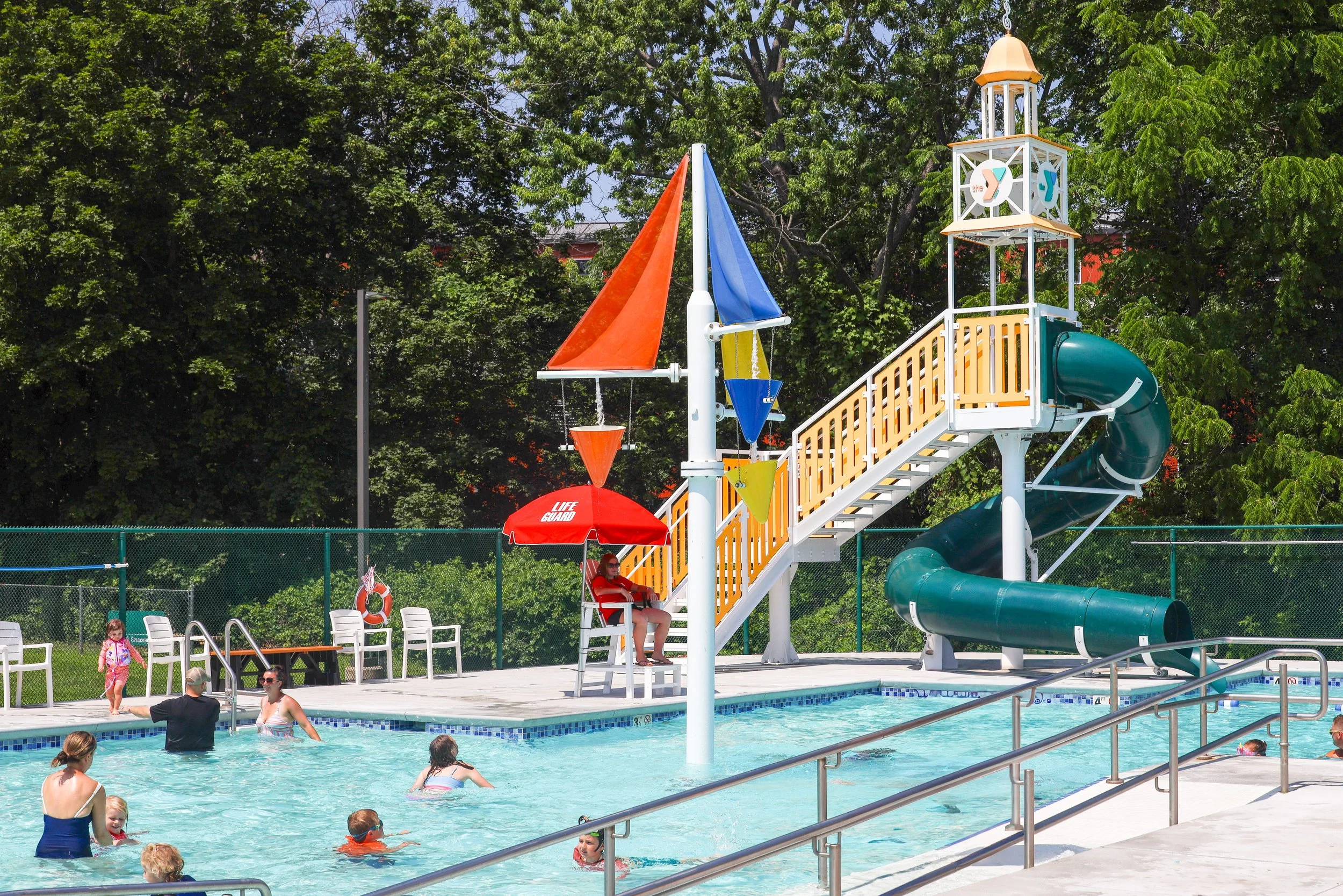 Children and adults enjoying a public pool with a waterslide and water play structure, surrounded by green trees.