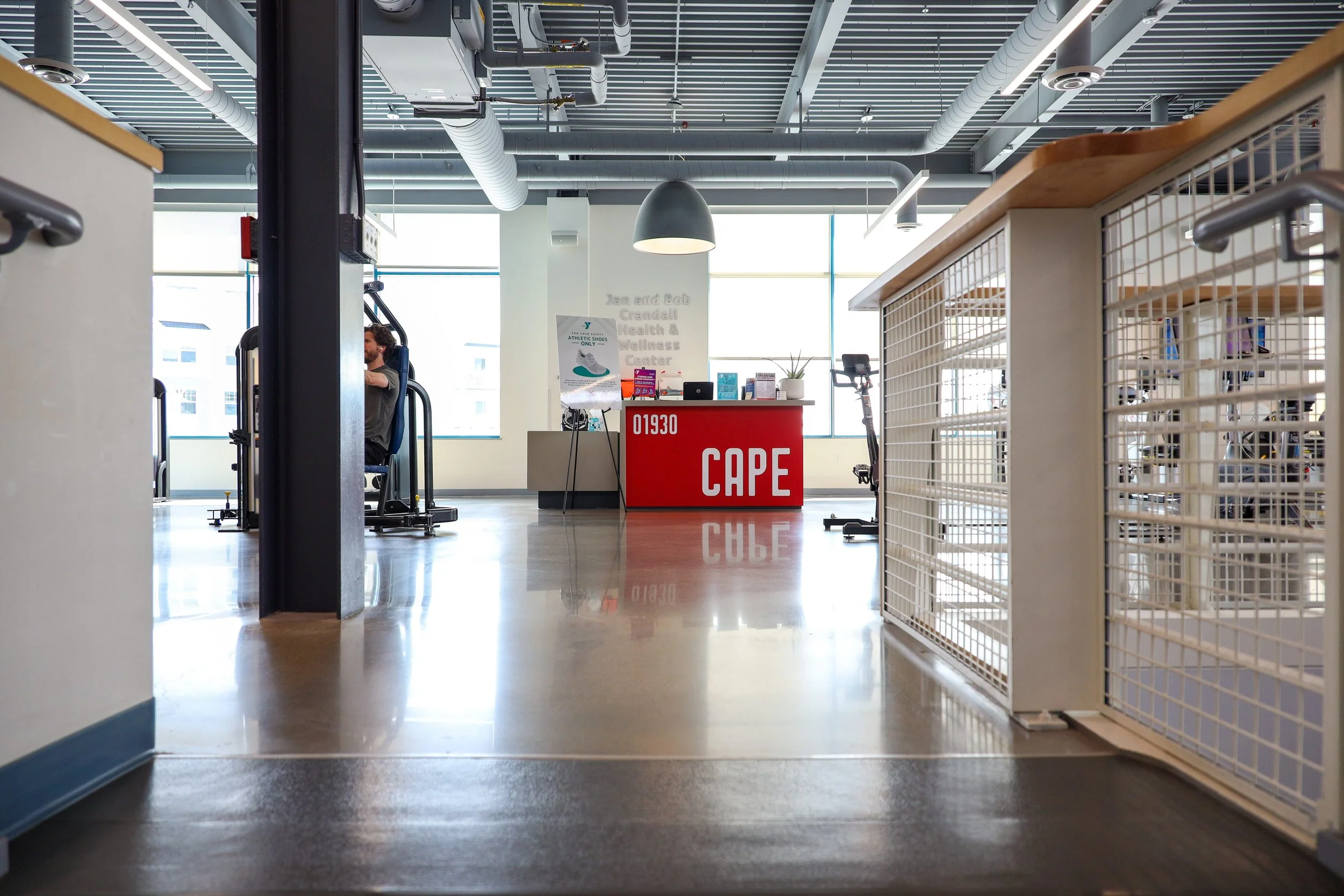 Interior of a fitness center with workout equipment, a red reception desk labeled 'CAPE,' and people exercising in the background.