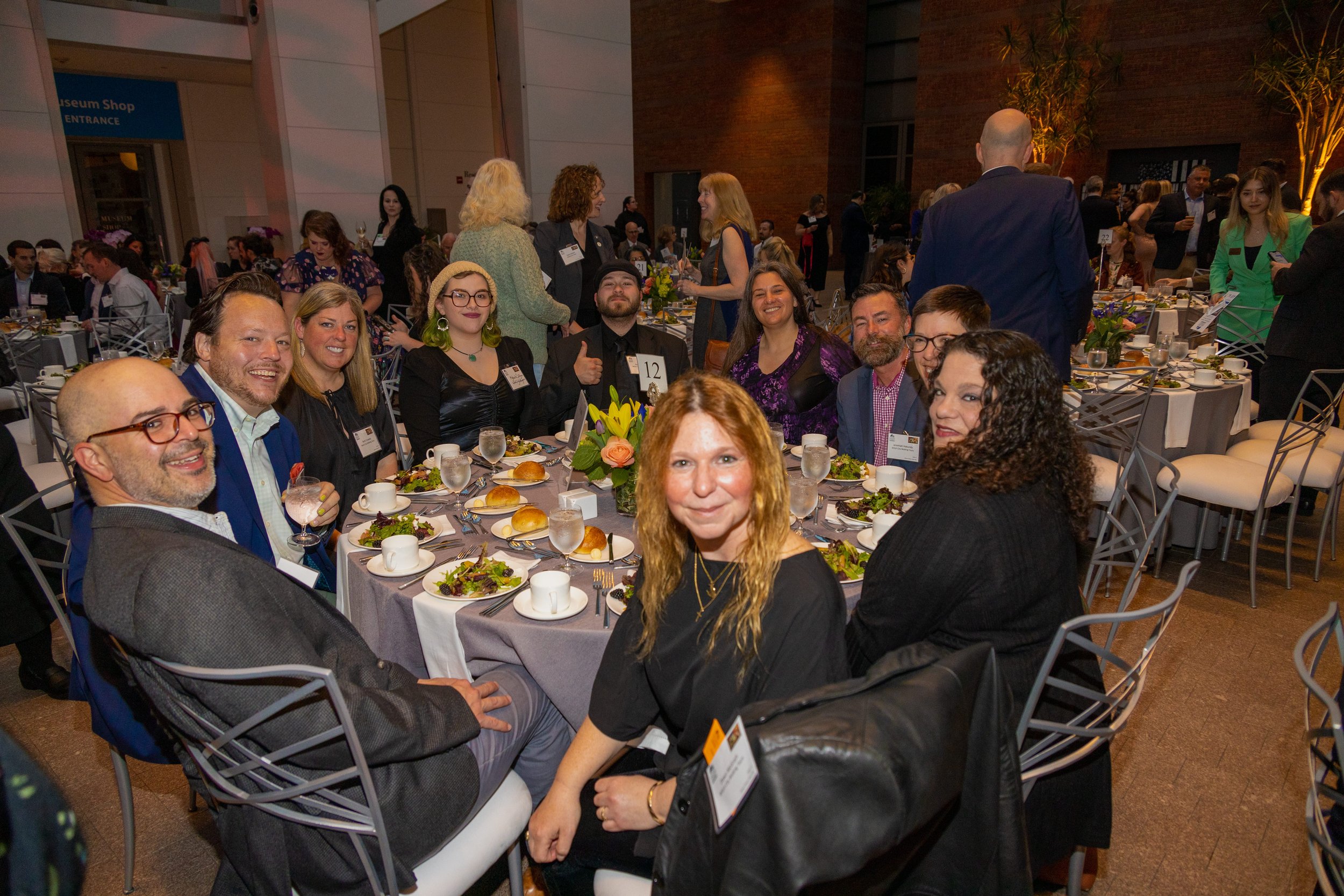 Group of people sitting around a round dinner table at a formal event, smiling, with plates of salad, bread rolls, and glasses of water, in a large, well-lit banquet hall.