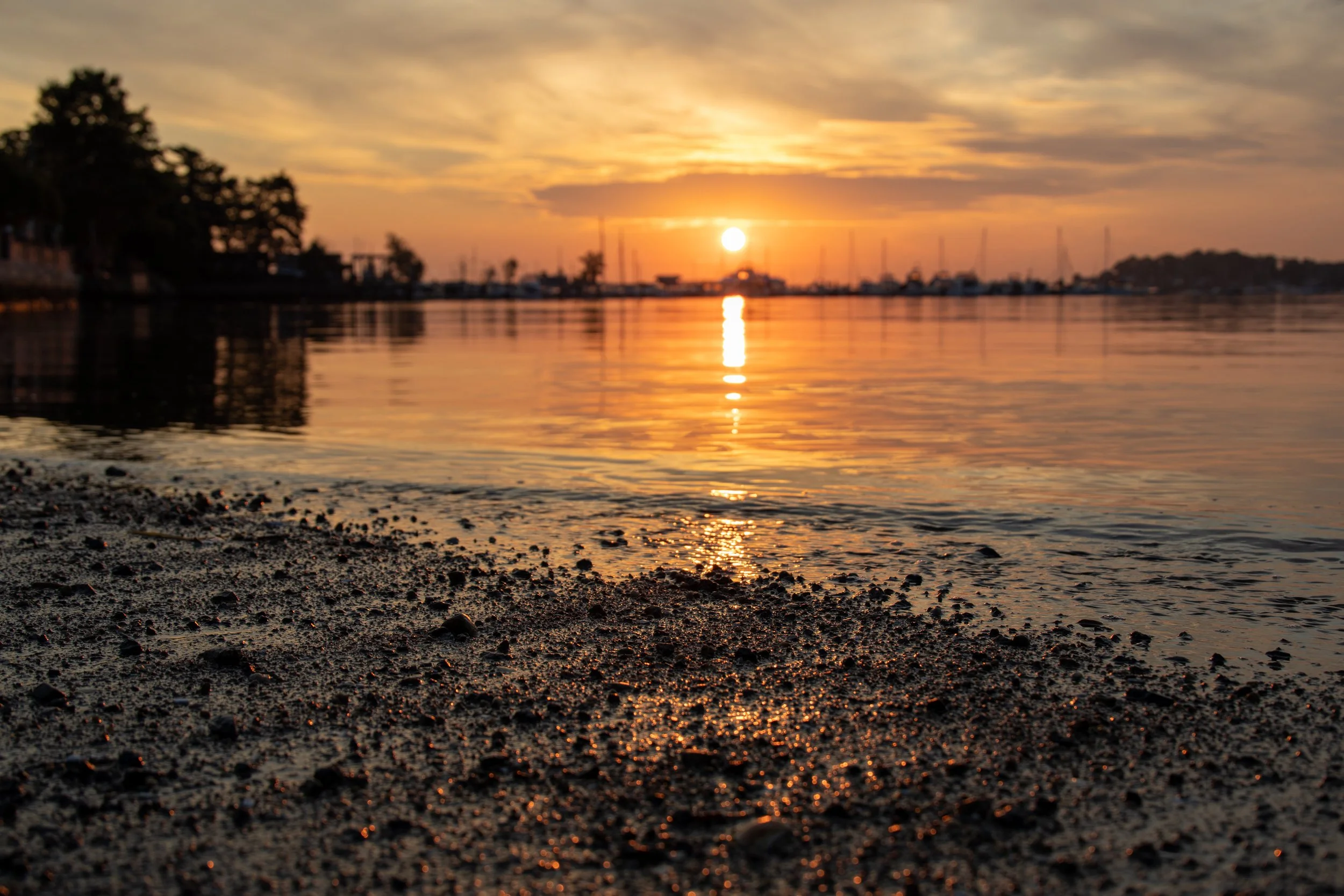 Sunset over a calm body of water with a sandy beach in the foreground and sailboats in the distance.