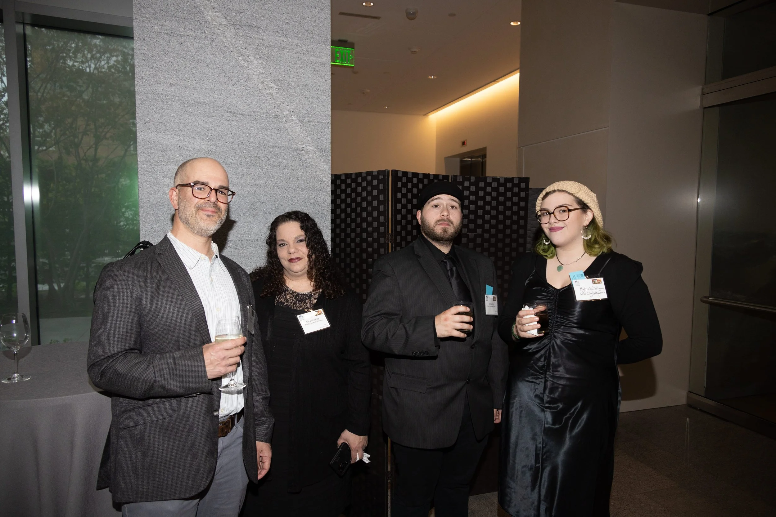 Group of four adults at a social event, holding drinks, standing indoors near a grey wall and large window, with name tags visible on two individuals.