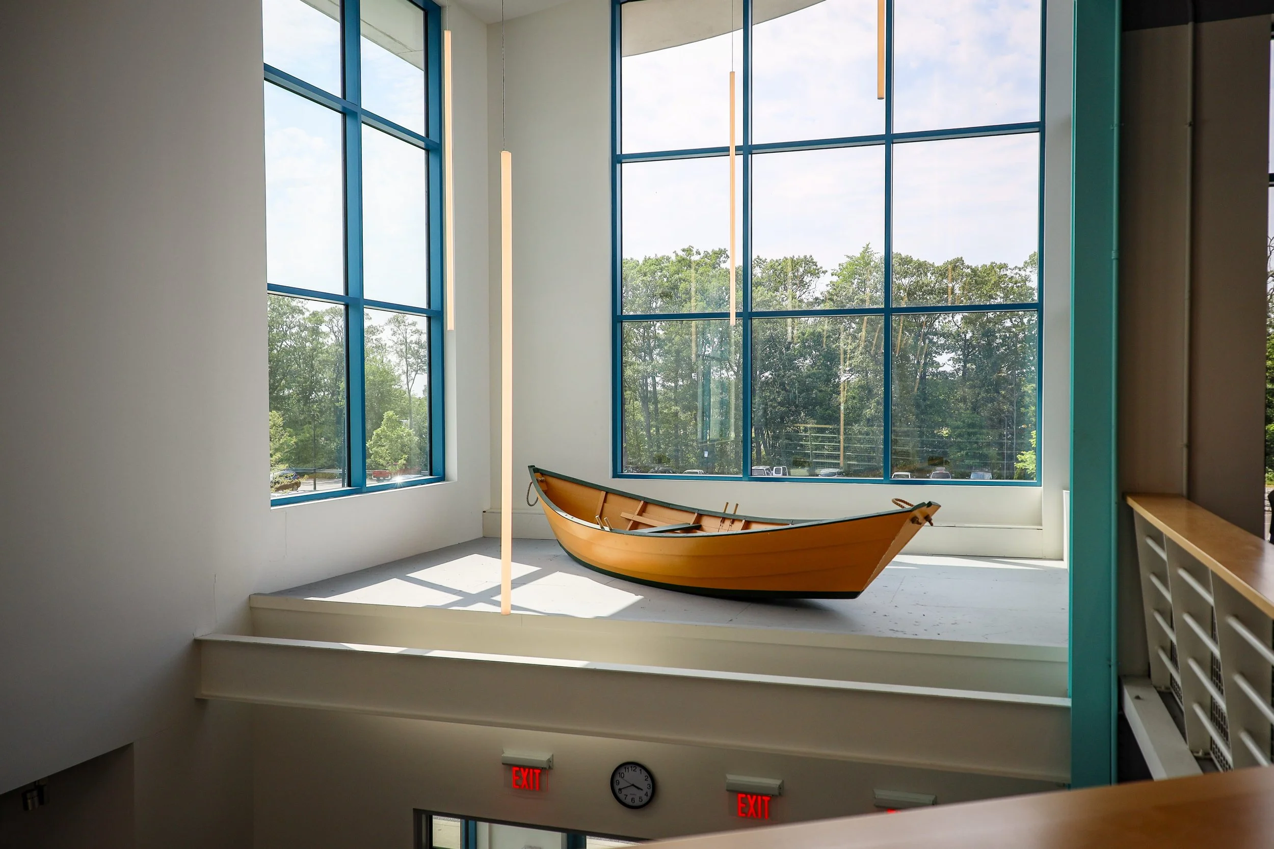 Interior of a building with large windows, a wooden boat displayed on a platform, and vertical hanging lights.