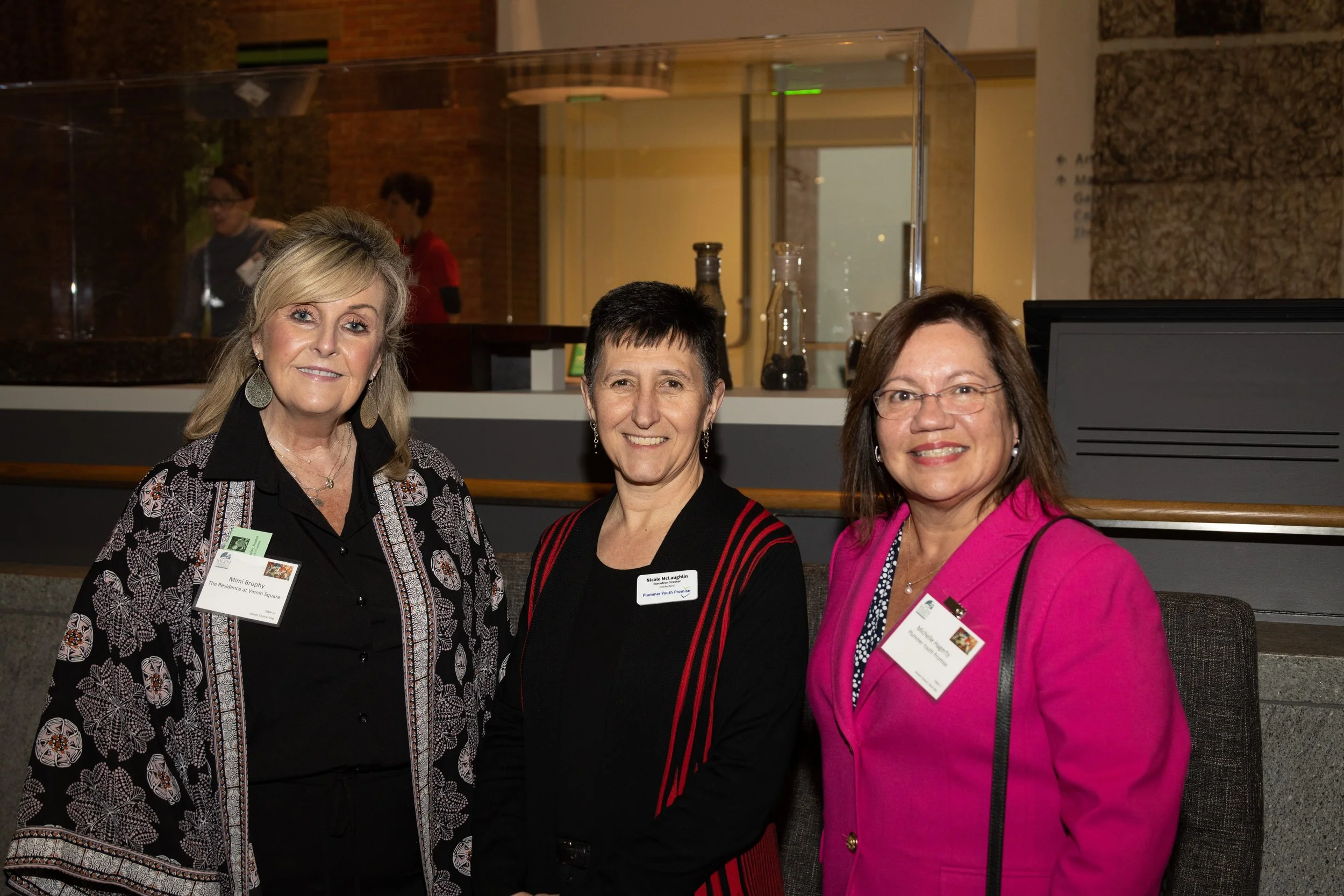 Three women standing together at an event, smiling at the camera, wearing name tags. The woman on the left has blond hair and is dressed in a black and patterned jacket. The woman in the middle has short dark hair and is wearing a black blazer with red stripes. The woman on the right has shoulder-length brown hair, glasses, and is dressed in a bright pink blazer.
