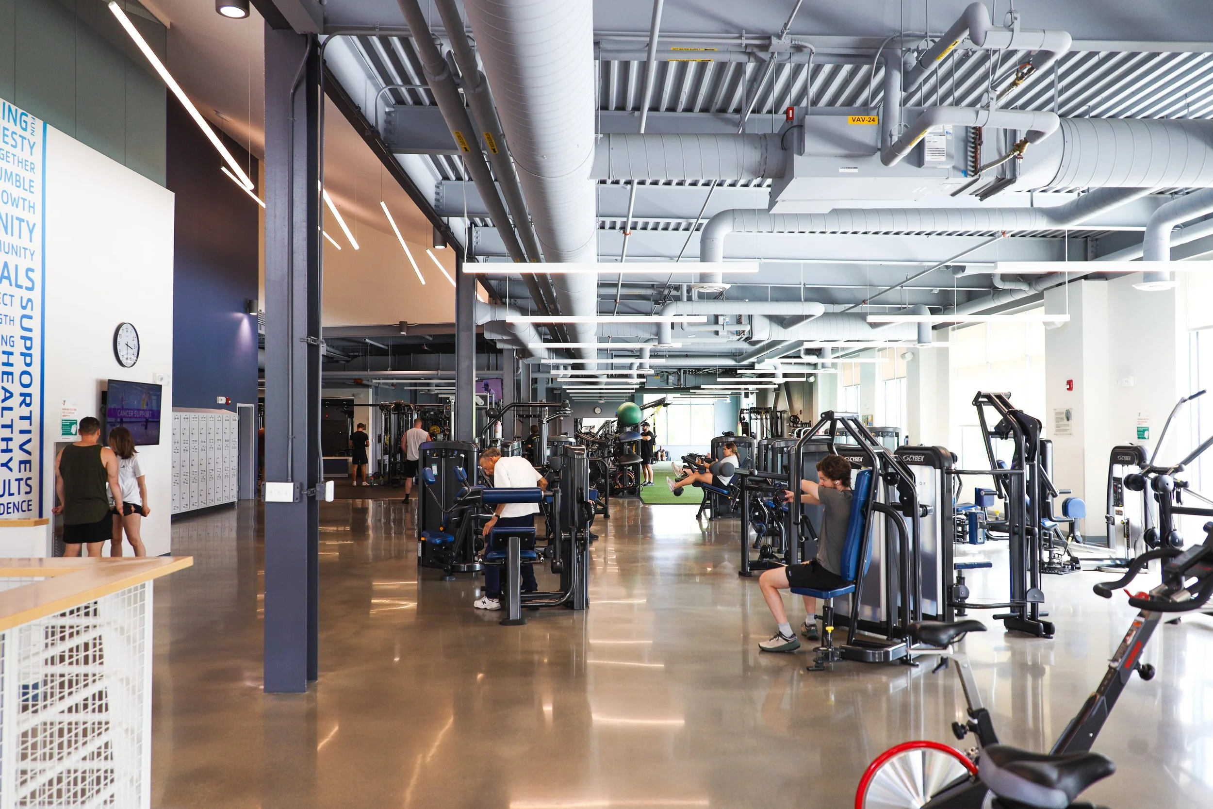 Inside a modern gym with various workout equipment and people exercising, bright natural light coming through large windows.