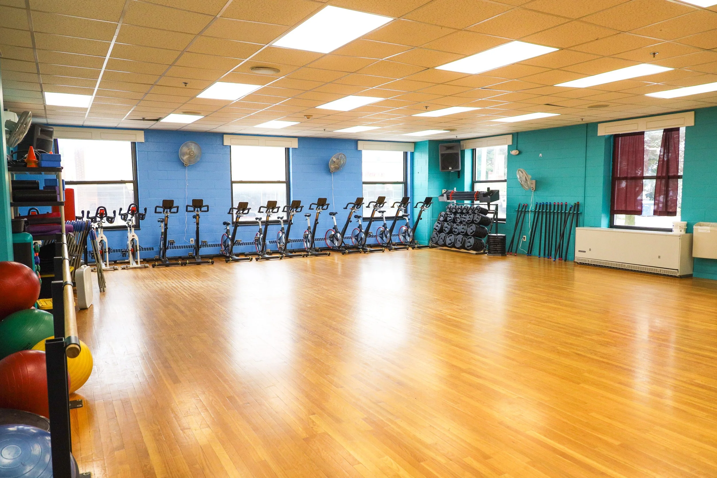 Empty workout room with exercise bikes, yoga mats, and fitness equipment along a bright blue wall with windows.