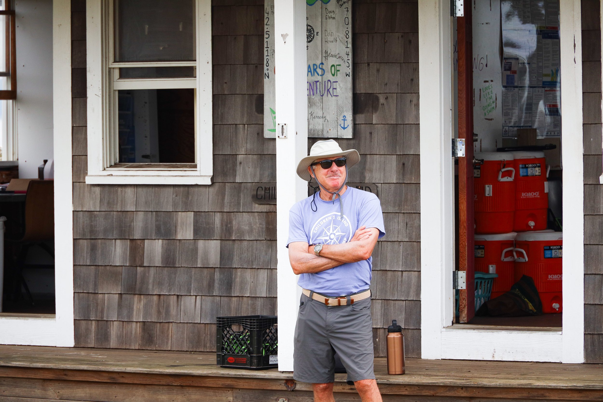 An older man with a tan hat, sunglasses, and a blue t-shirt with a compass design stands on a wooden deck outside a small building, crossing his arms. Behind him are two open doors revealing storage items, including red coolers, and a window with a white frame.