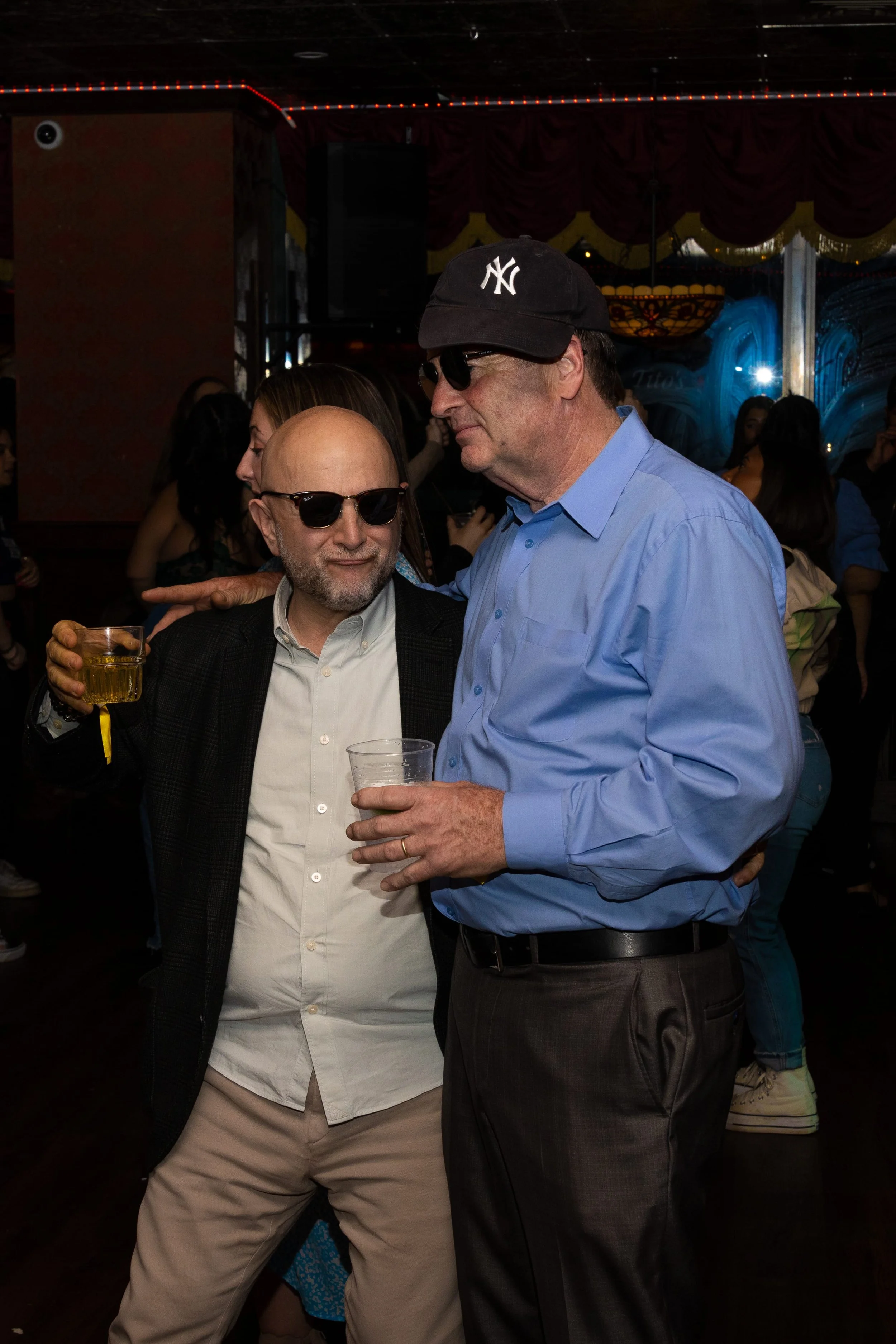 Two men wearing sunglasses and holding drinks are conversing closely at a social gathering or party, with a woman in the background and colorful decorations on the wall.