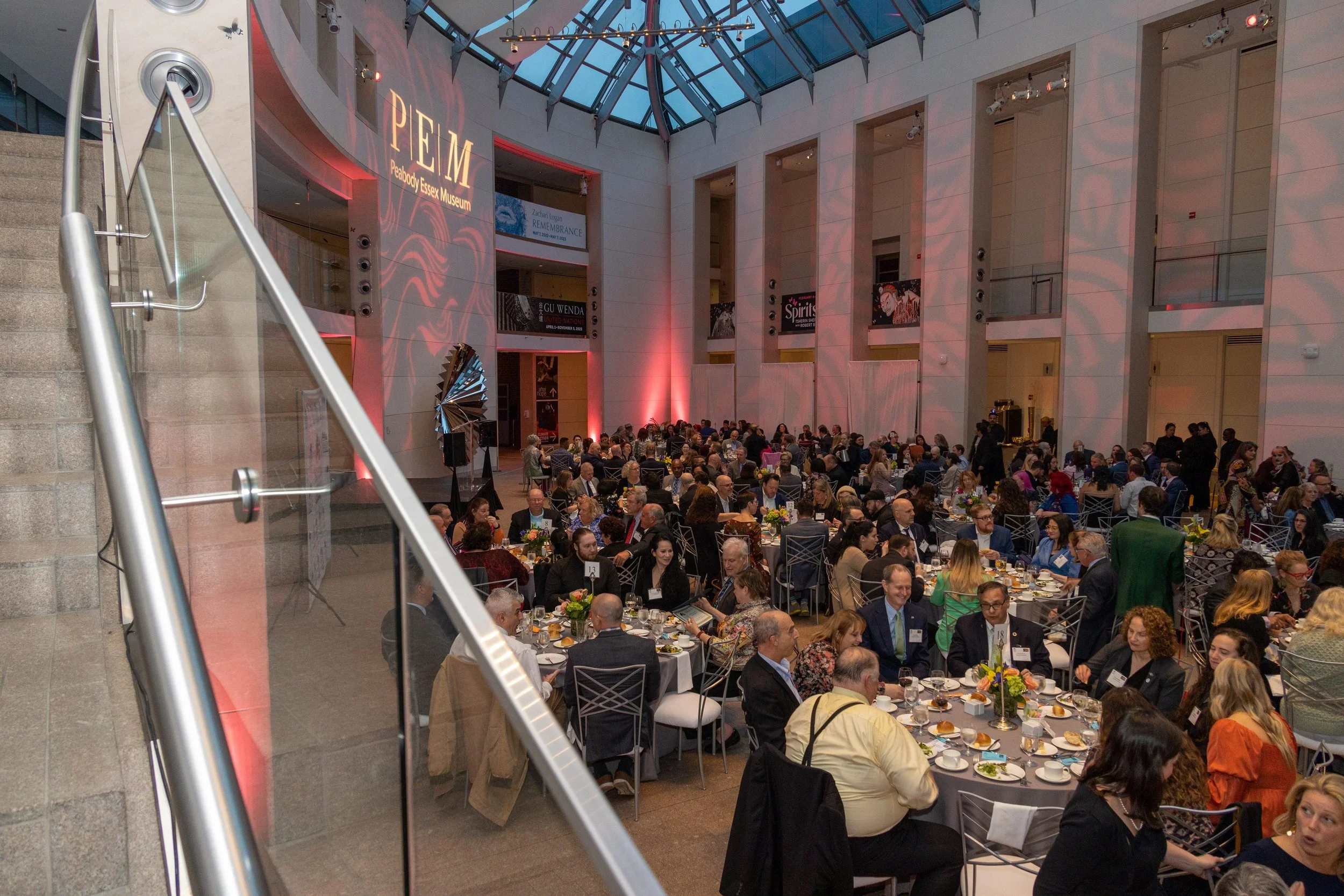 A large indoor event at Peabody Essex Museum with many round tables set for a formal dinner, filled with attendees in business attire, in a spacious, modern atrium with high glass ceilings and banners on the walls.
