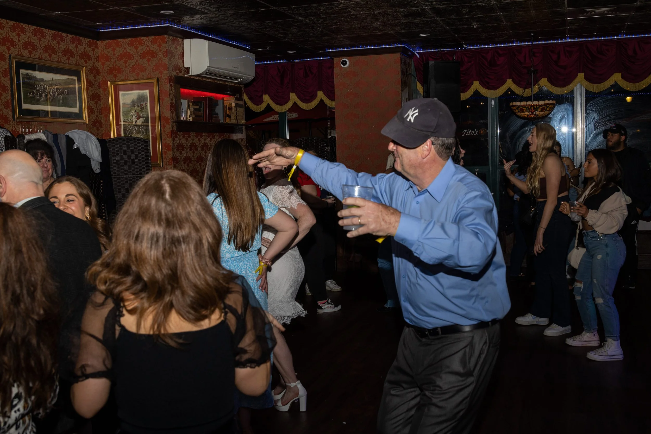 A man dancing at a party or nightclub with a drink in hand, surrounded by other people socializing and dancing indoors.
