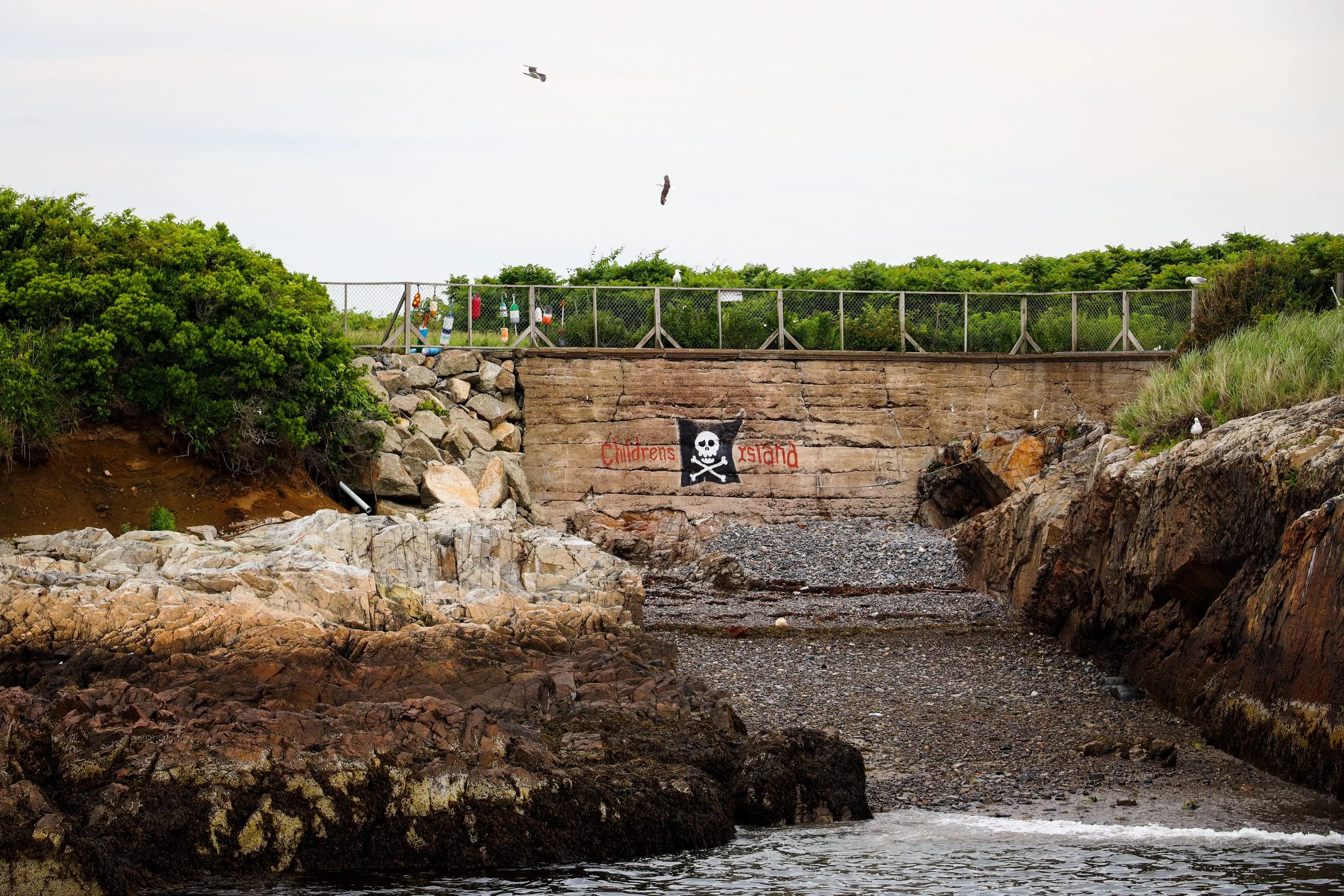 Rocky shoreline withgraffiti that reads 'Children's Island' and a skull and crossbones symbol, with a wooded area and a fence on top in the background.