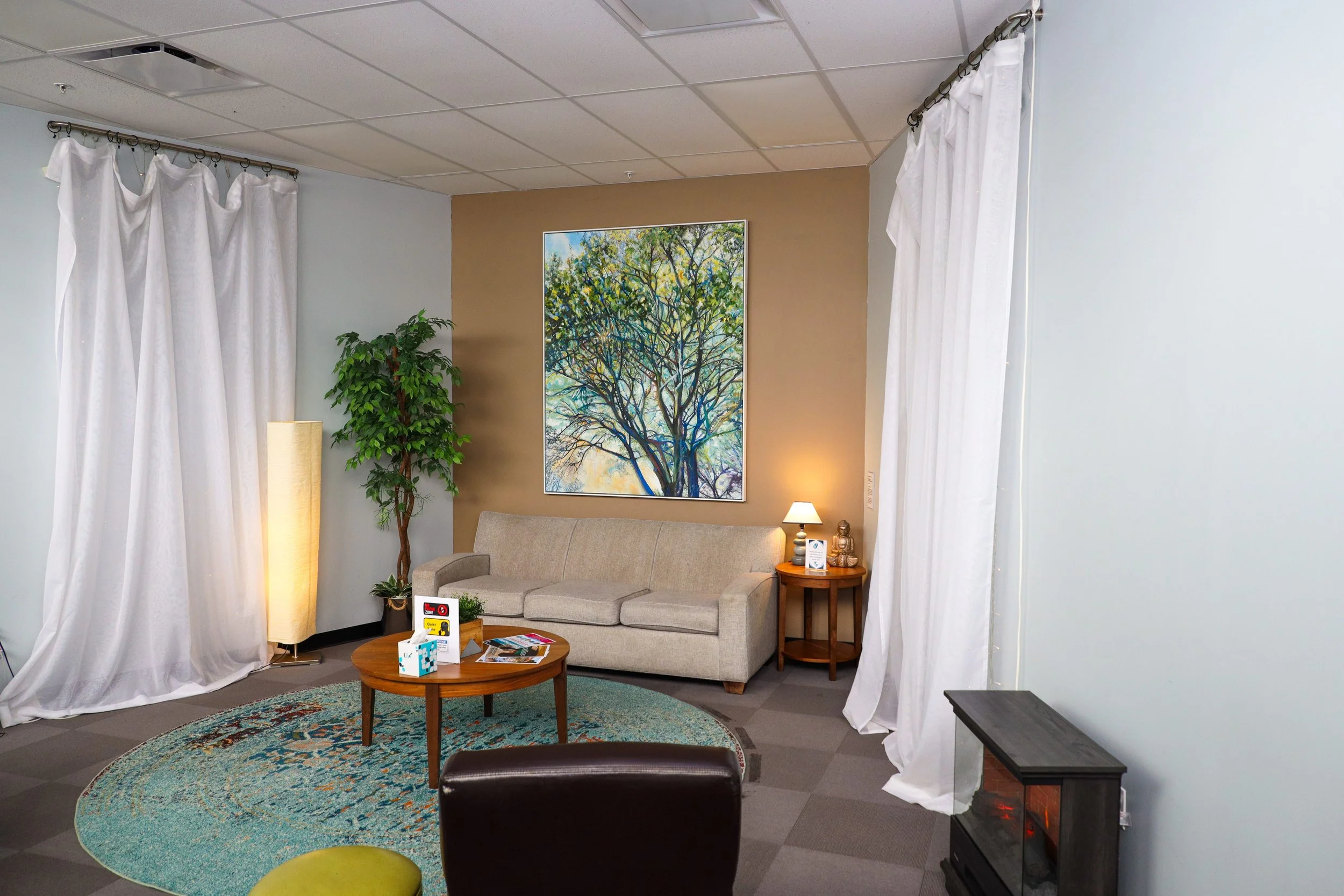 Comfortable waiting room with beige sofa, side table with lamp and Buddha statue, framed colorful tree painting on wall, green potted plant, floor lamp, white curtains, round rug, and electric fireplace.