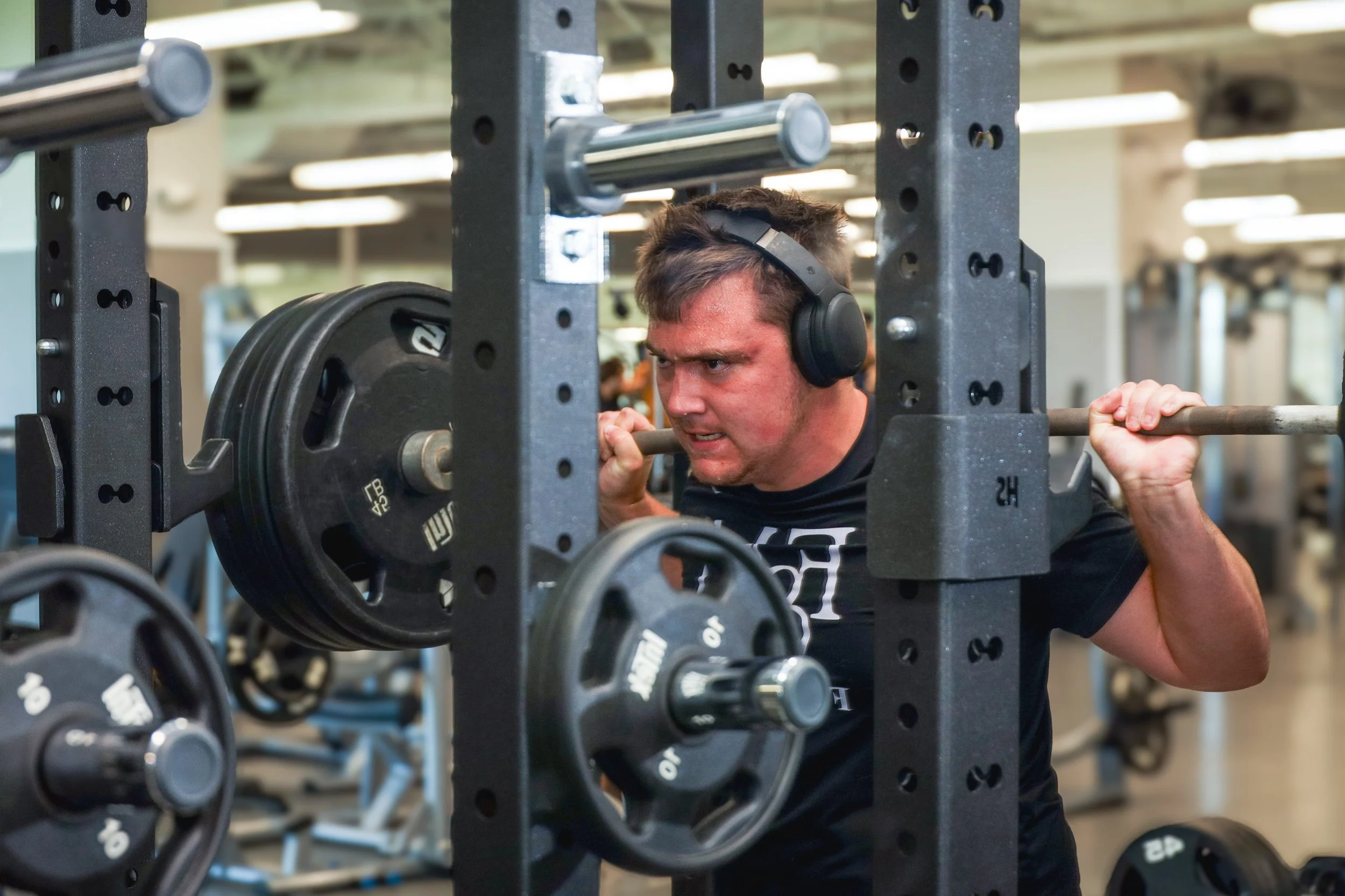 A man wearing headphones lifts a barbell behind a squat rack in a gym, displaying a determined facial expression.