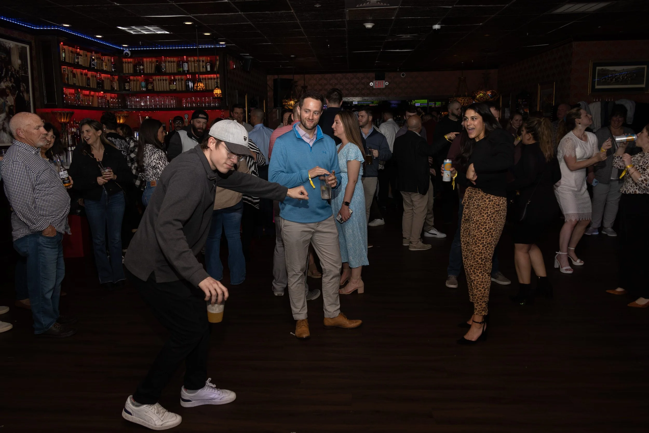 People dancing and socializing at a lively bar or nightclub with dim lighting and a decorated bar counter in the background.
