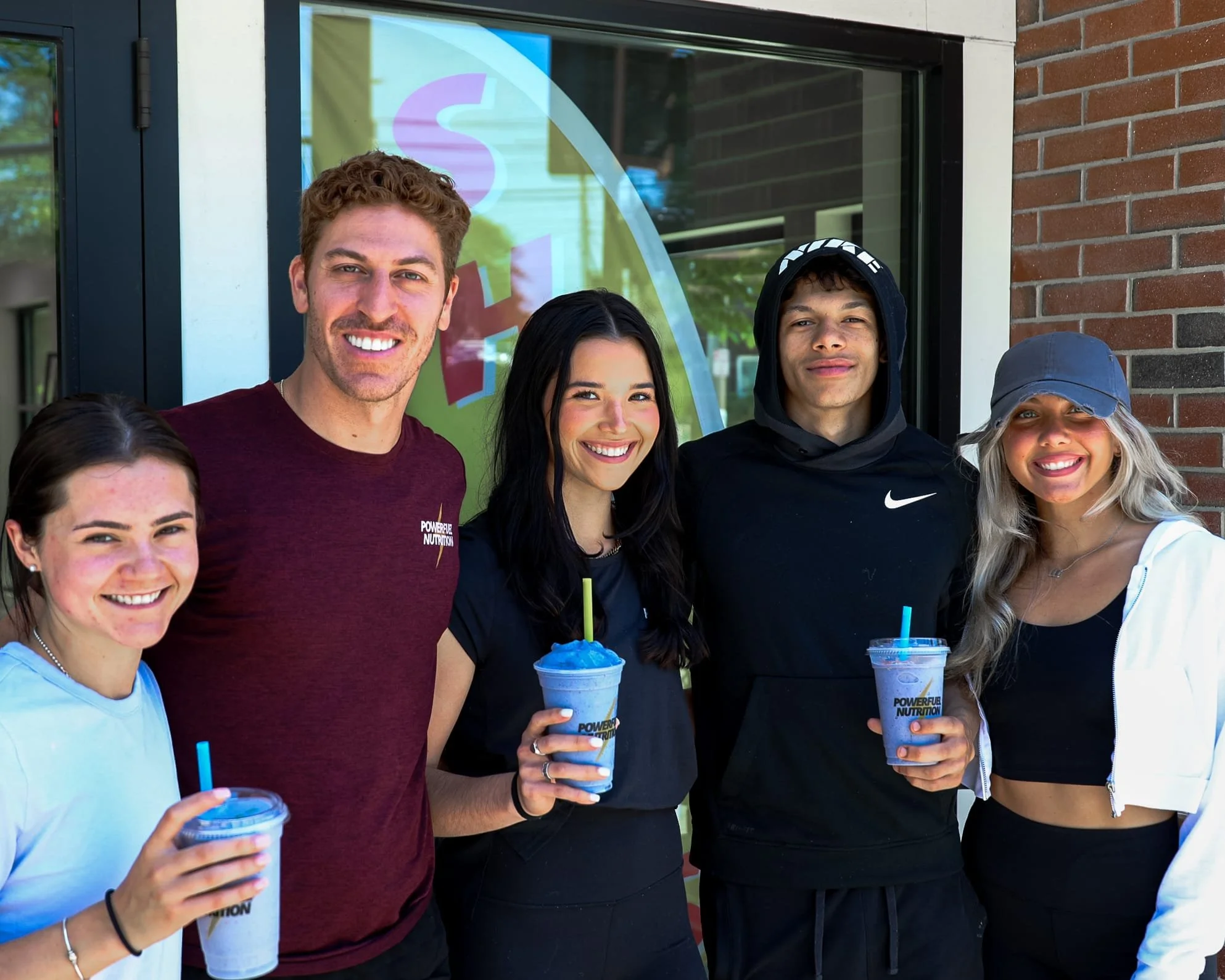 Group of five young adults standing outdoors, smiling, holding colorful smoothies, in front of a glass door with a colorful logo on it, a brick building to the right.