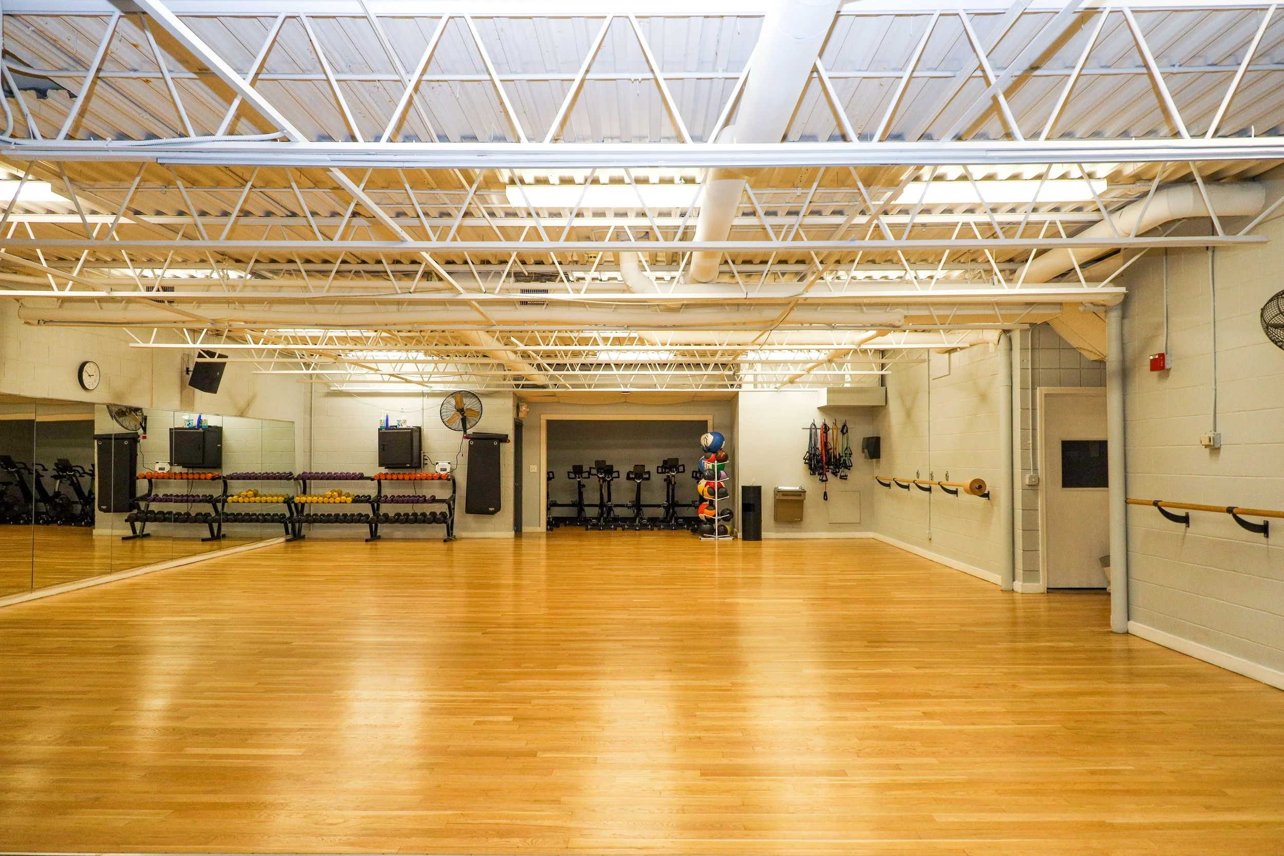 Empty indoor fitness studio with wooden floors, large mirror wall, and workout equipment including weights, bikes, and resistance bands.