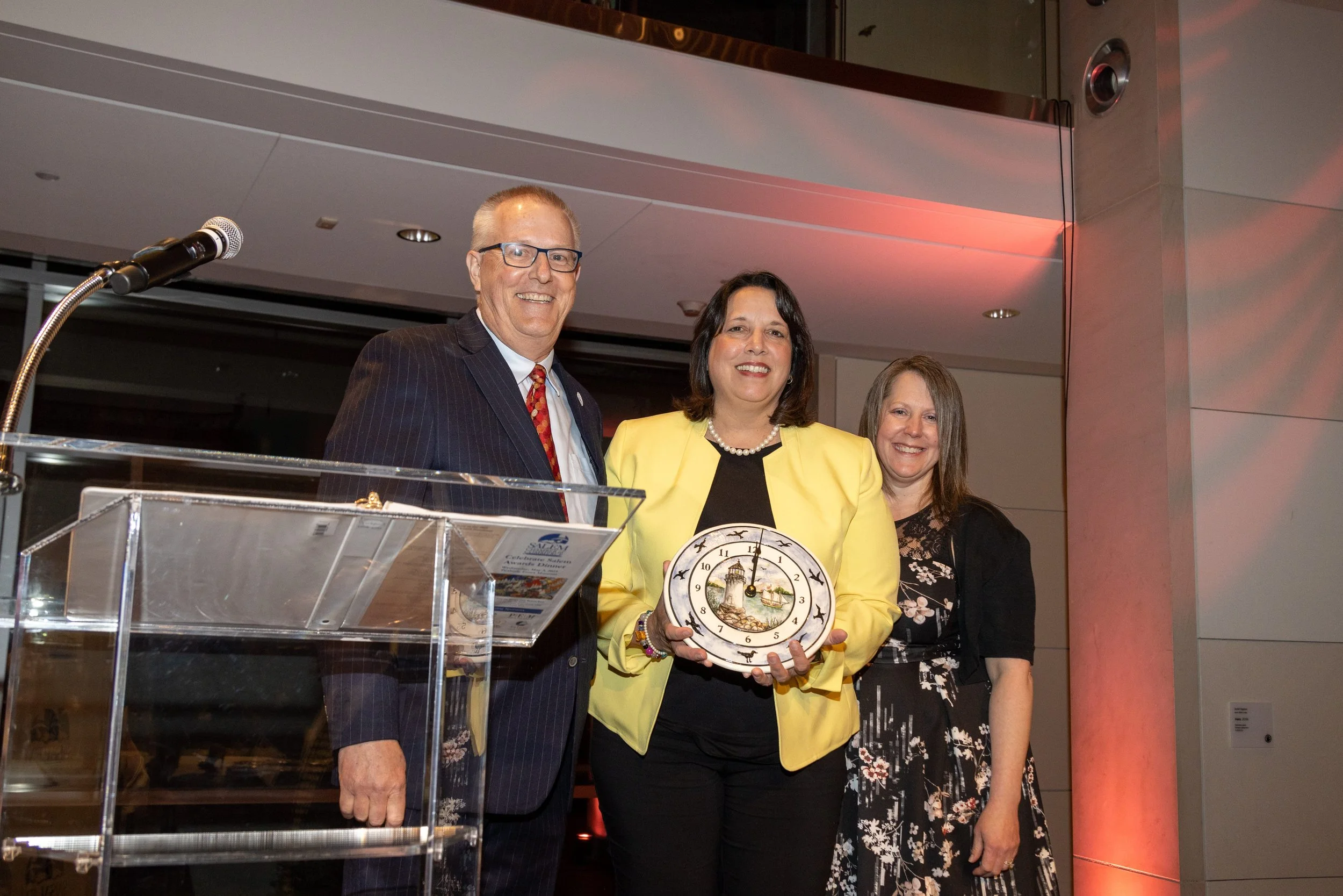 Three people standing indoors, one woman holding a clock, all smiling, at a formal event or gathering.