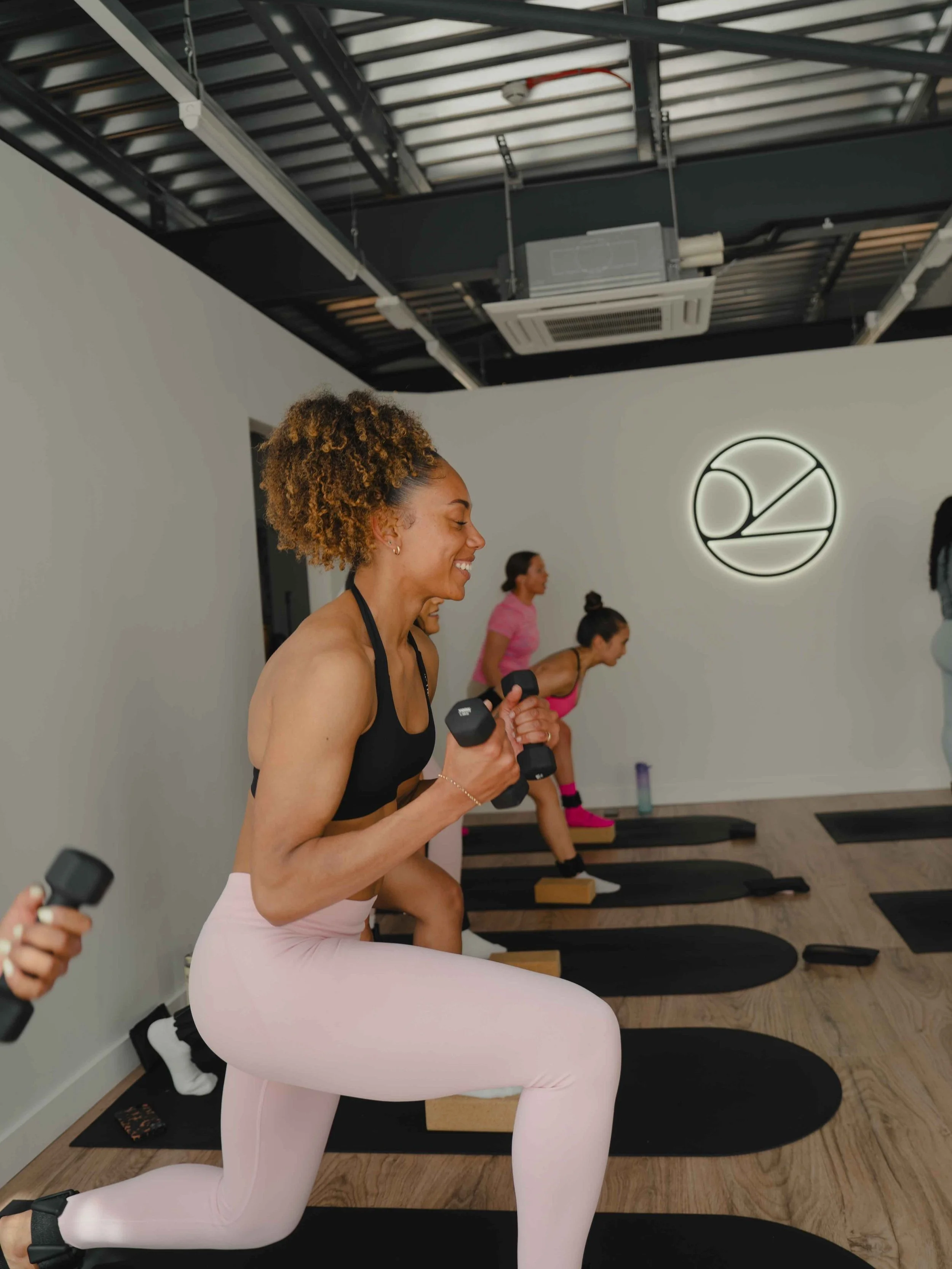 A group of women exercising in a fitness studio, lifting dumbbells and lunging on mats. The woman in the foreground is smiling and wearing a black sports bra and light pink leggings.