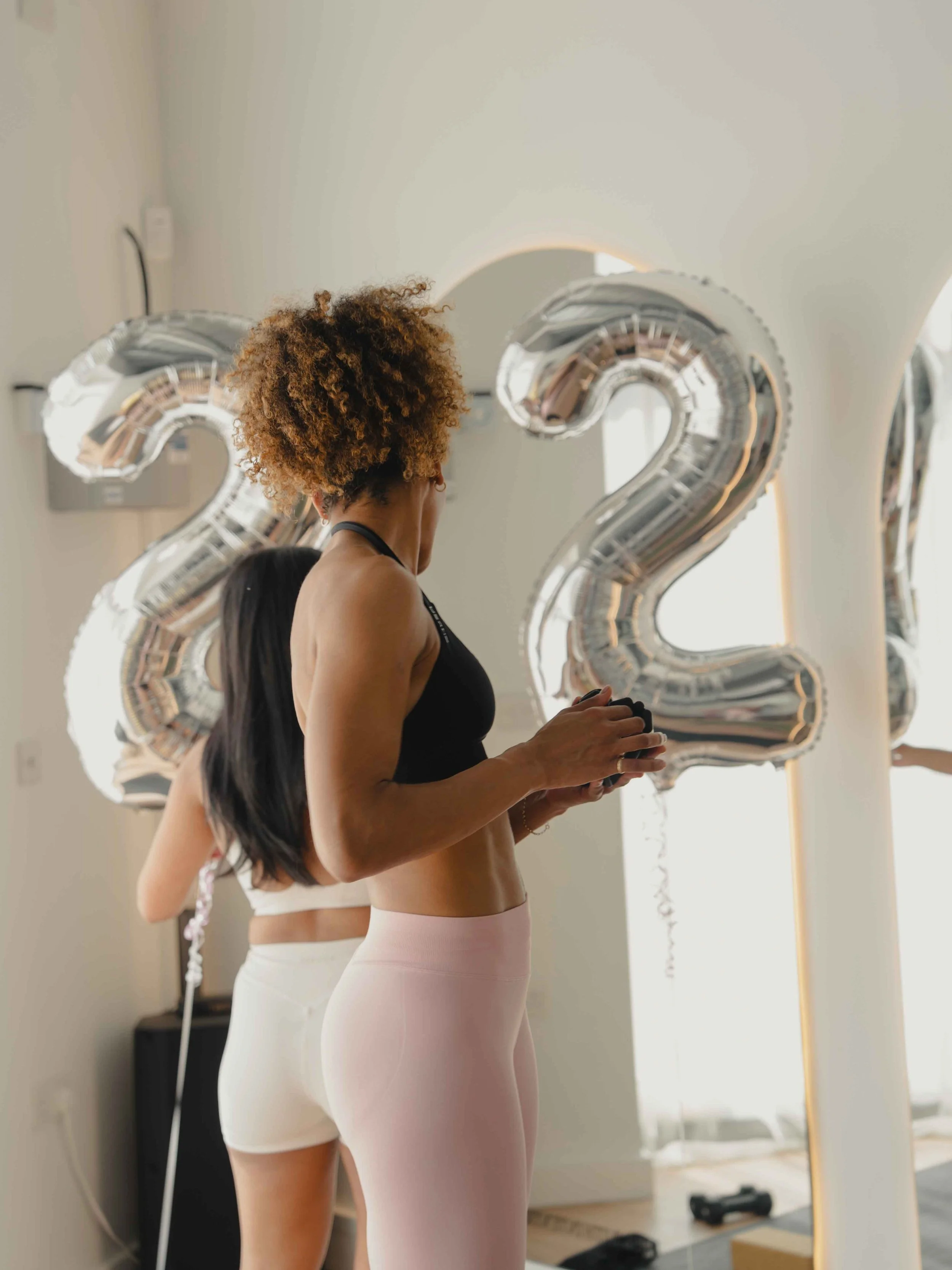 Two women in workout clothes standing indoors with large silver balloon numbers '2' and '2' in the background.