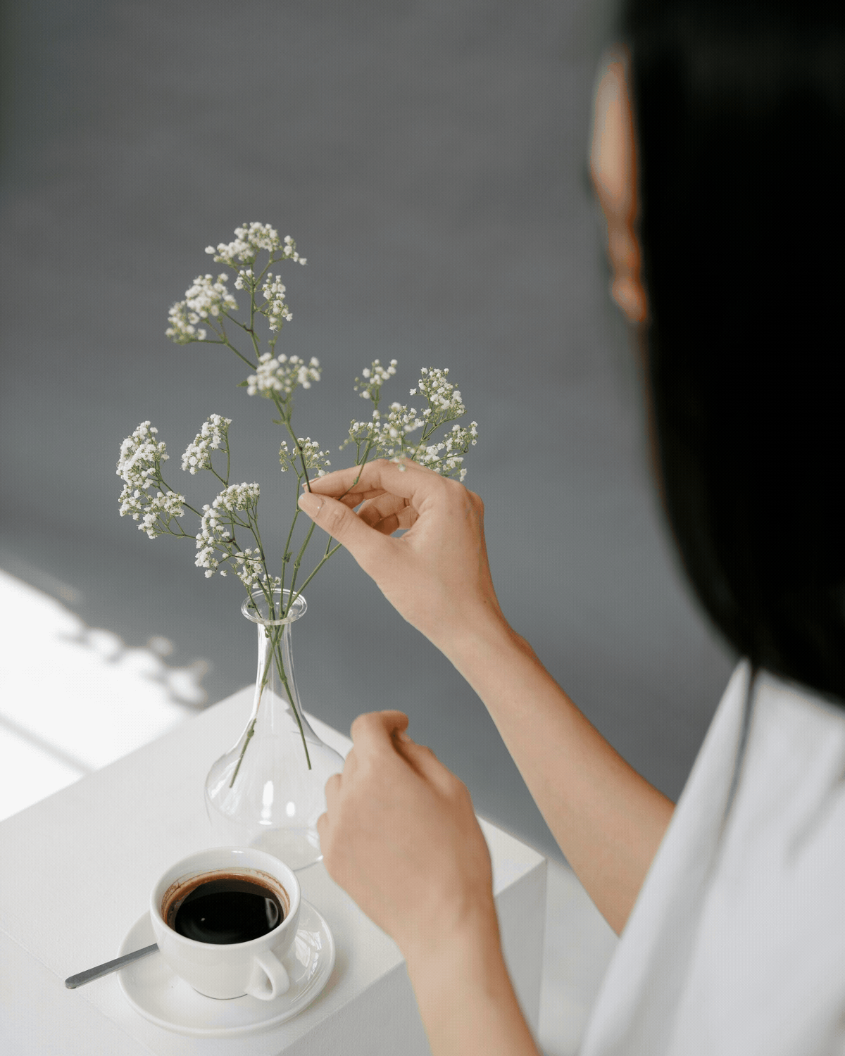 A person arranging white baby’s breath flowers in a glass vase, with a cup of black coffee and a spoon on a white table.