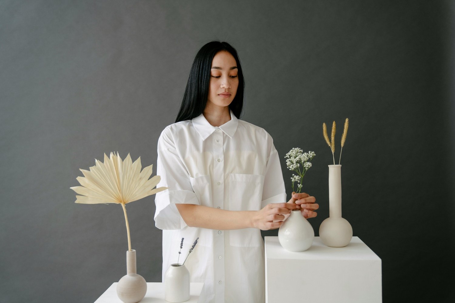 Woman with long black hair arranging white flowers and grasses in white ceramic vases on display tables in a minimalistic setting.