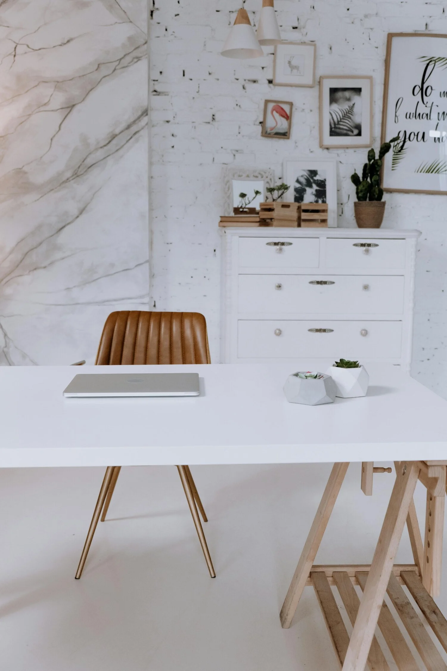 Modern workspace featuring a white table with a closed silver laptop and two small geometric planters with succulents, a brown leather chair, a white dresser with decorative items and framed art on a white brick wall, and a gallery of framed art and quotes.