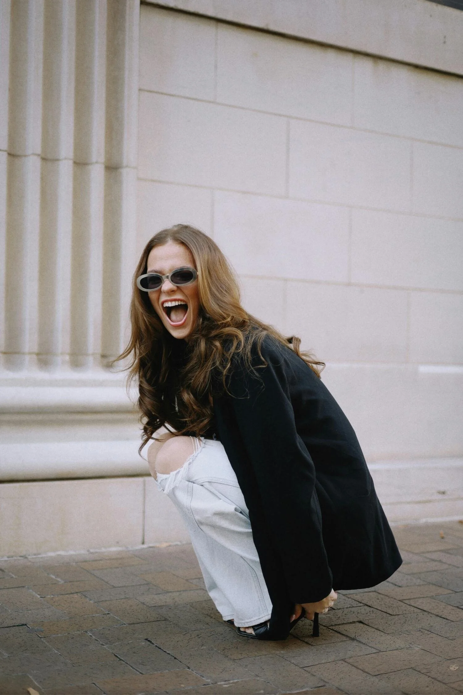 A woman with long brown hair wearing sunglasses, a black jacket, and white ripped jeans, crouching on the sidewalk near a building with stone walls, laughing and opening her mouth wide.