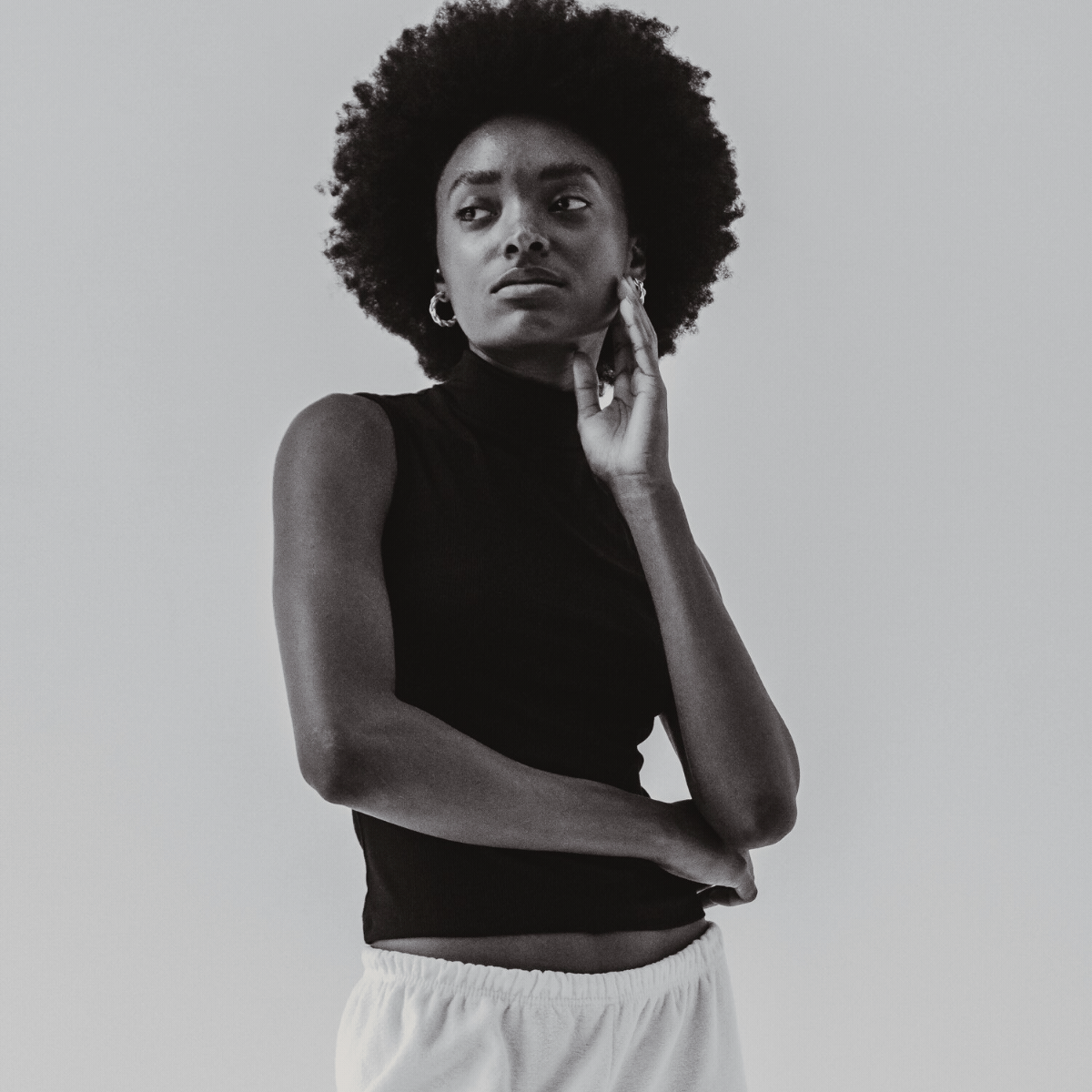 Black and white photograph of a young woman with curly hair, wearing a sleeveless black top and light-colored shorts, standing against a plain background with her hand gently touching her face.