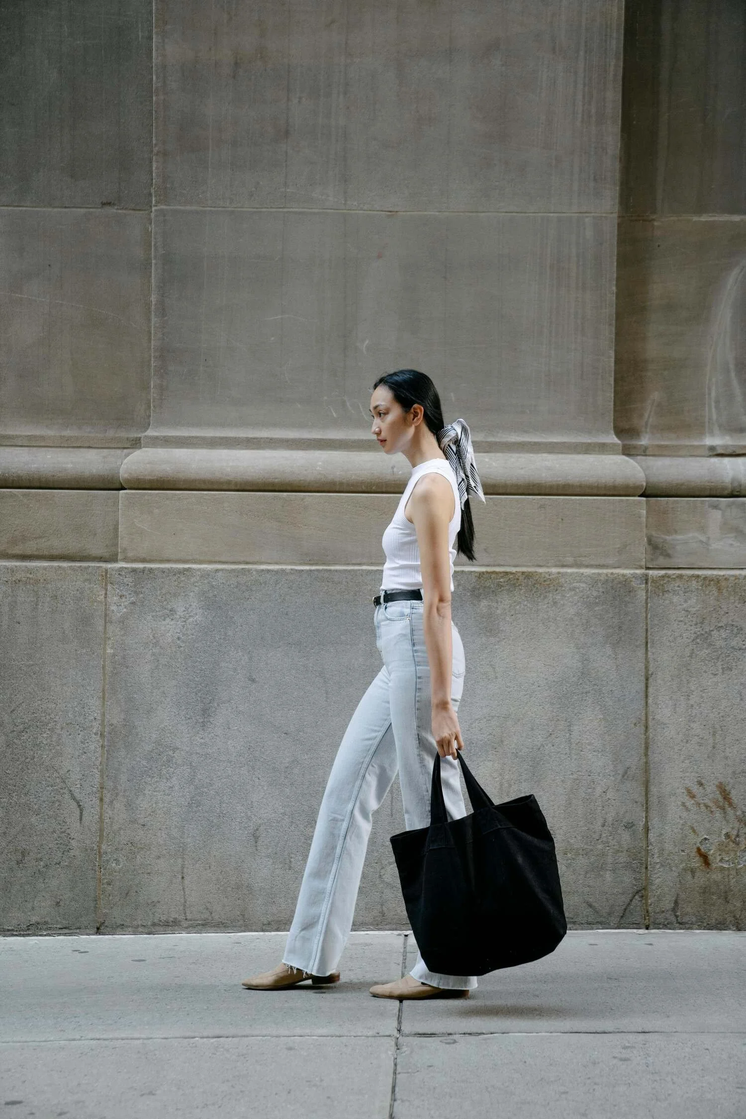 A woman walking on the sidewalk with a large black tote bag, wearing a white sleeveless top, light-colored jeans, and beige shoes, against a stone wall background.