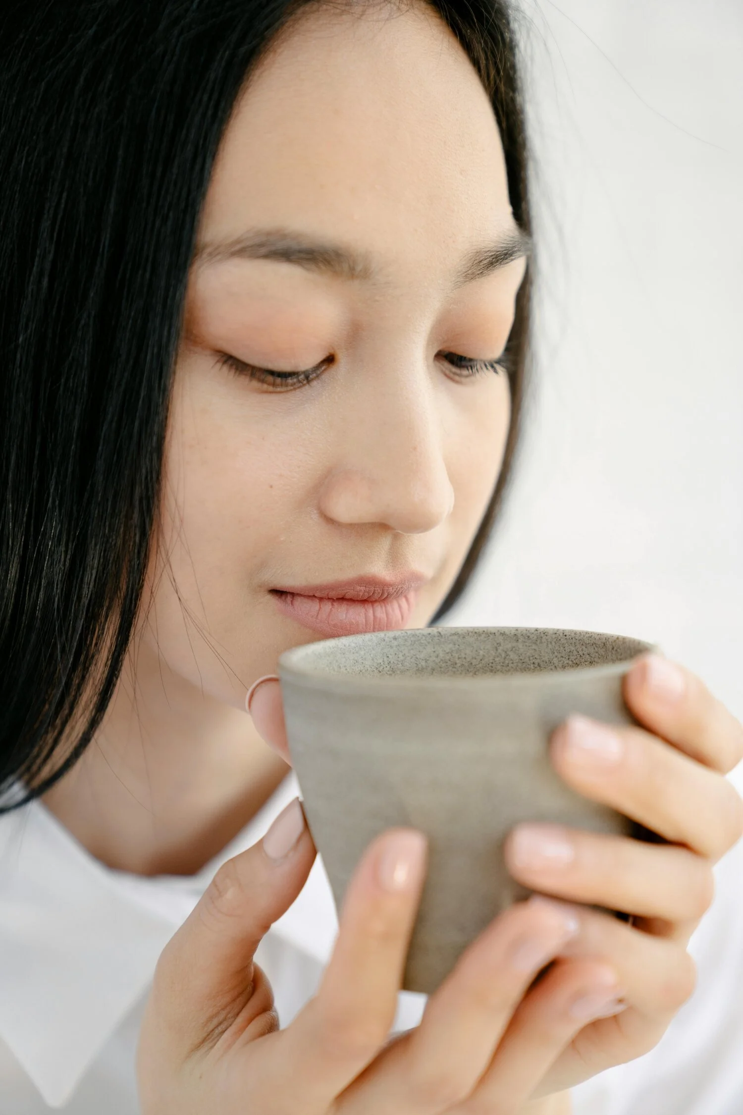 A woman with long dark hair holding a beige ceramic mug close to her face with her eyes closed, savoring the aroma of her drink.
