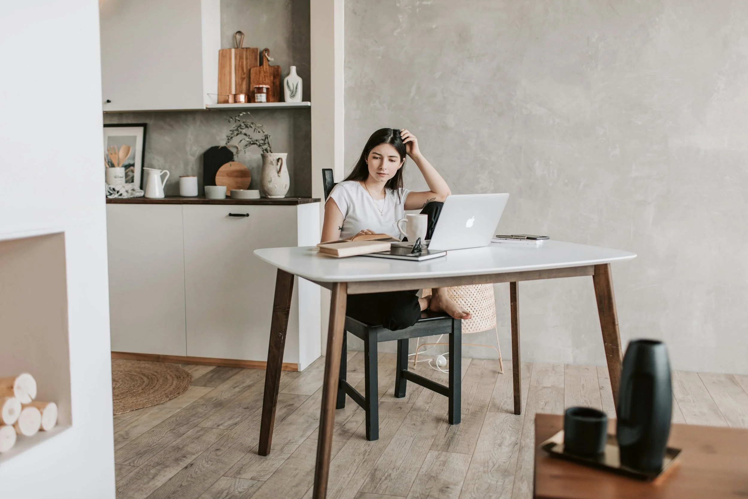 A woman sitting at a desk with a laptop, looking confused or distracted, in a modern, minimalist kitchen or dining area.