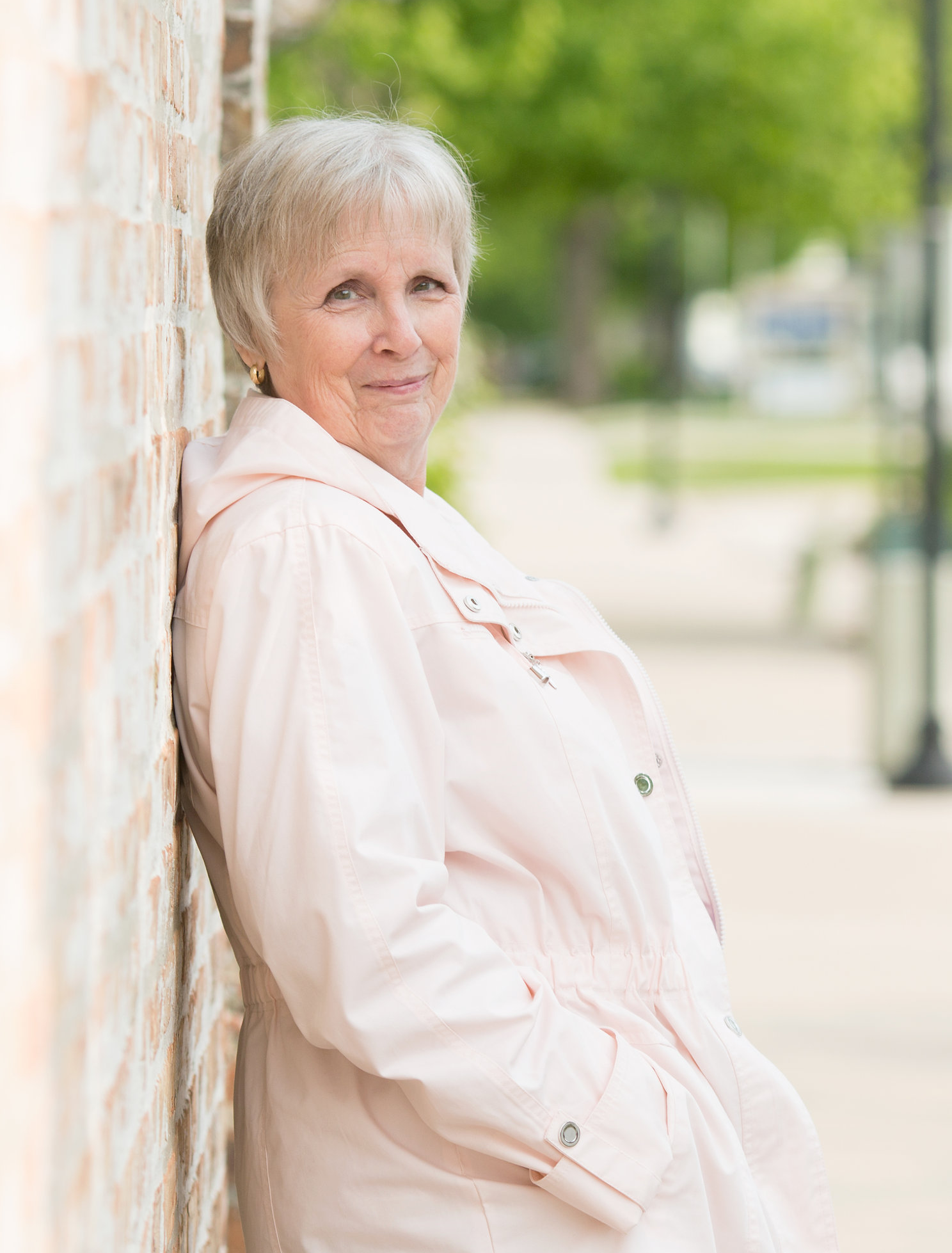 A woman with short hair leaning against a brick wall outdoors, wearing a light pink jacket, smiling softly, with greenery and blurred trees in the background.