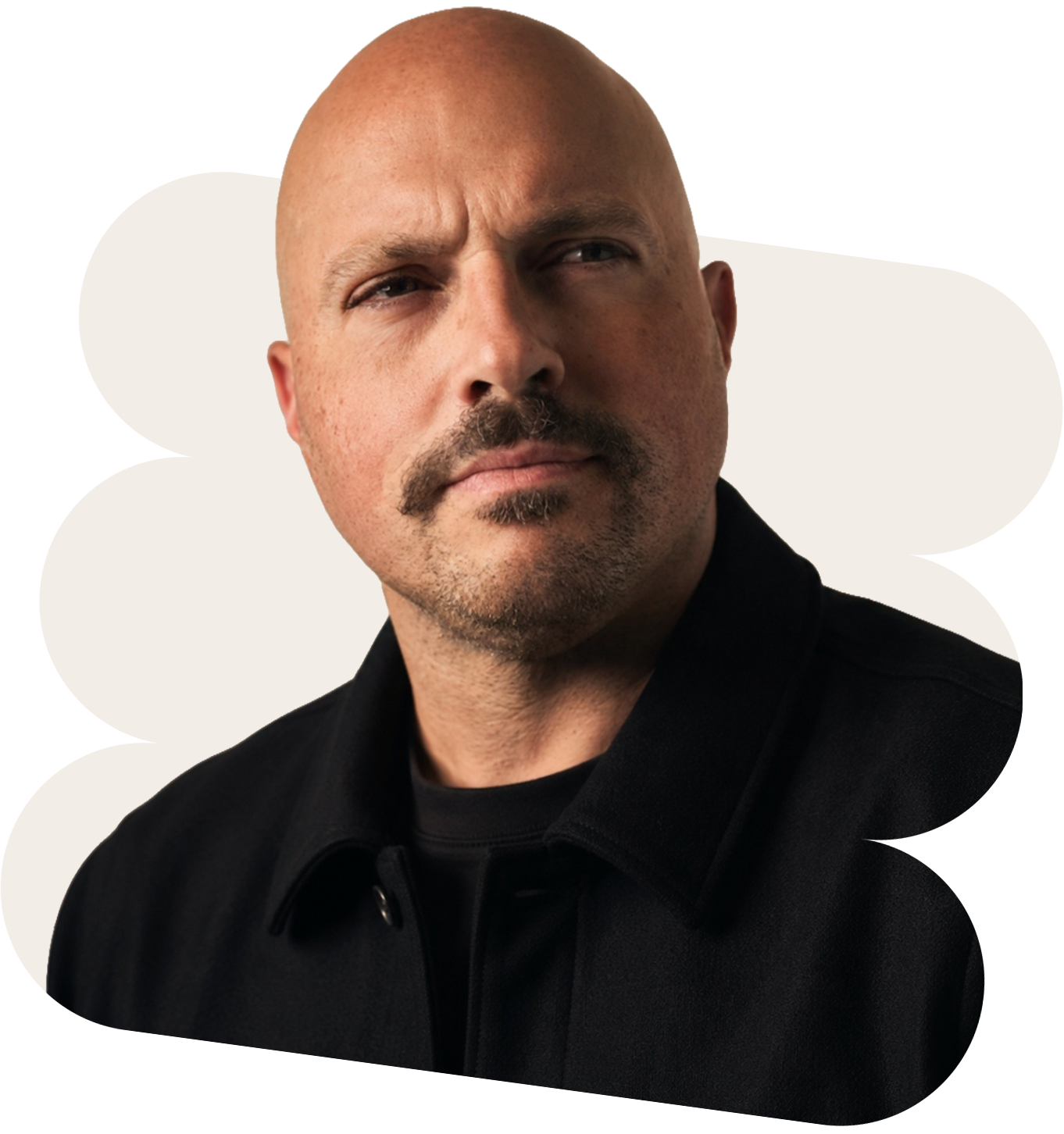A headshot of a serious expression man with a bald head, mustache, and goatee, wearing a black shirt with collar, looking to his right against a neutral background.