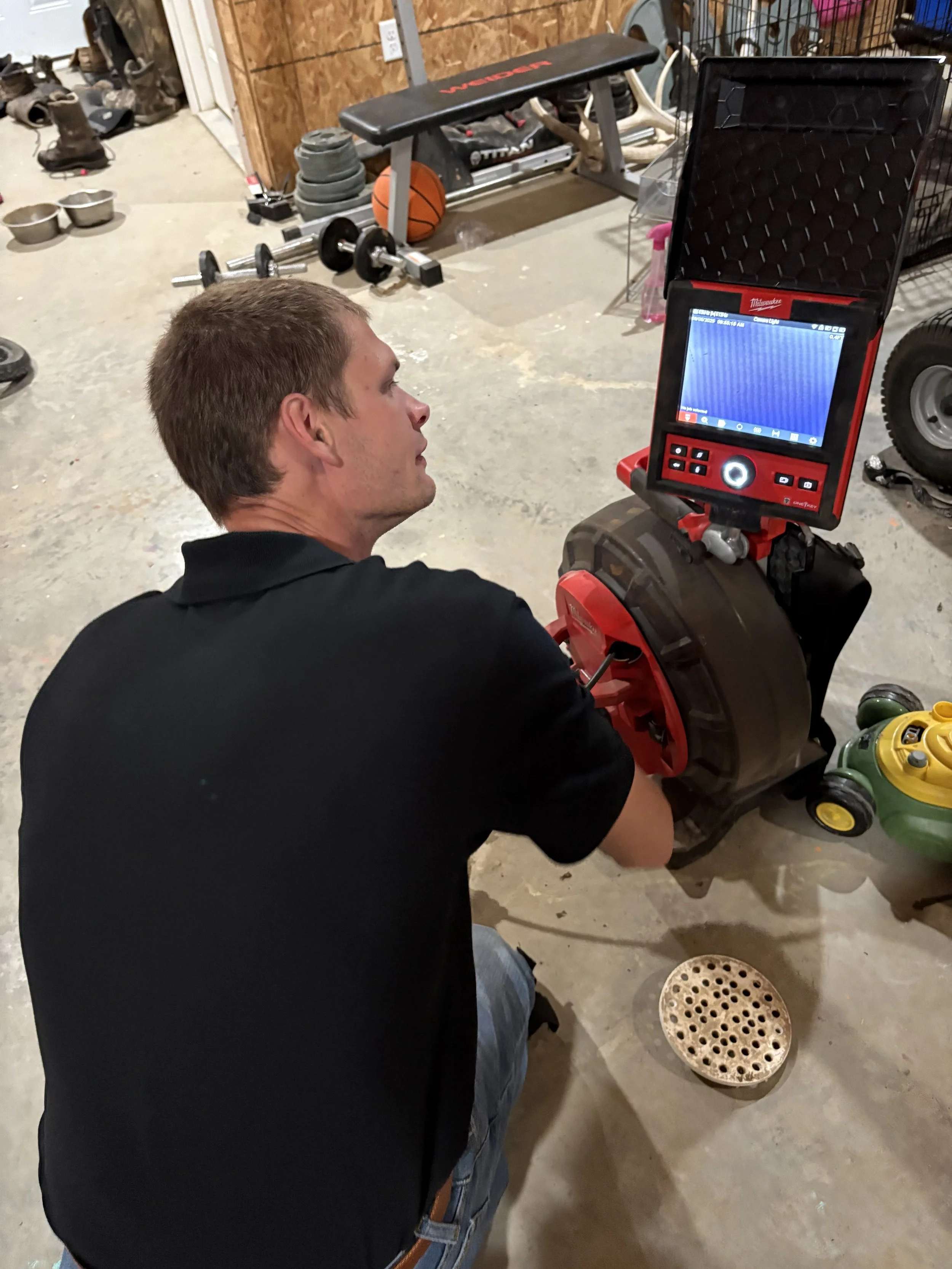A man is kneeling on the floor of a garage or workshop, working on a red and black device with a screen. The background shows various gym equipment, a basketball, and tools.